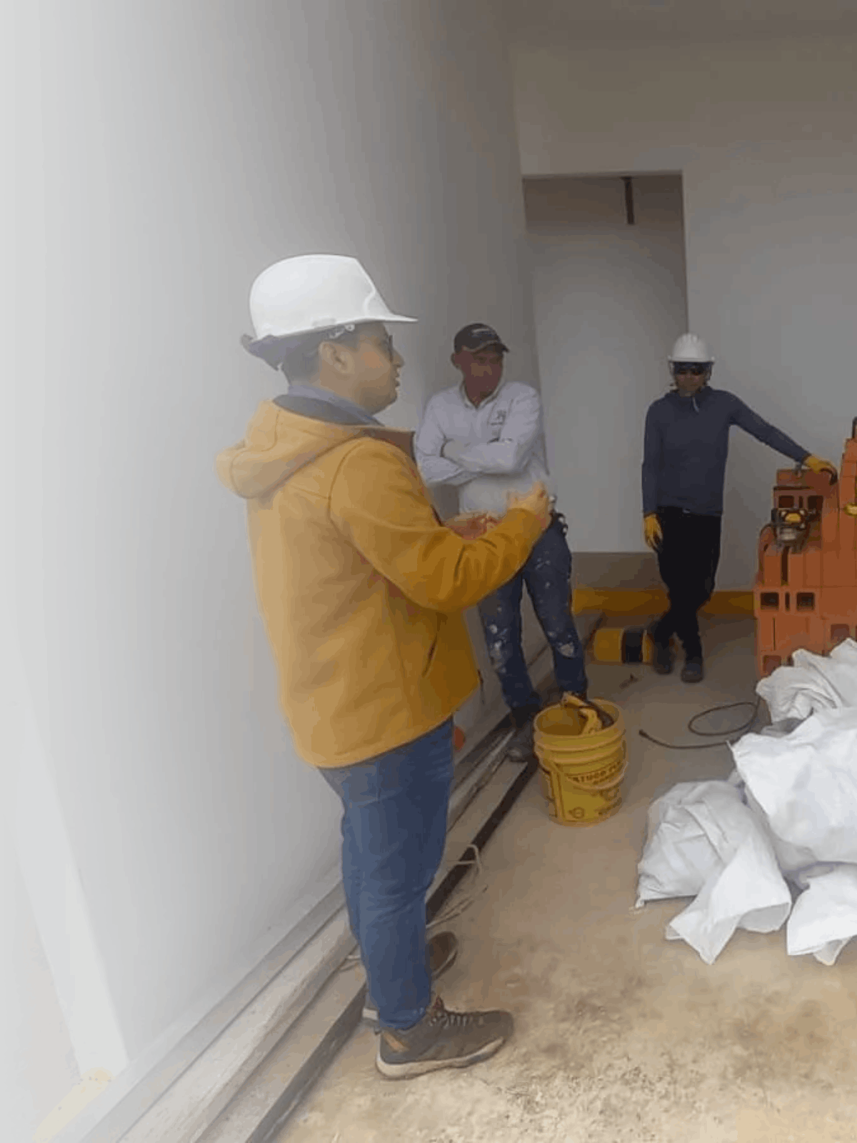 Tres trabajadores con cascos de seguridad en una construcción, uno con sudadera naranja, otro con camisa blanca y gafas, y otro con camiseta azul oscuro, junto a materiales y herramientas de construcción.