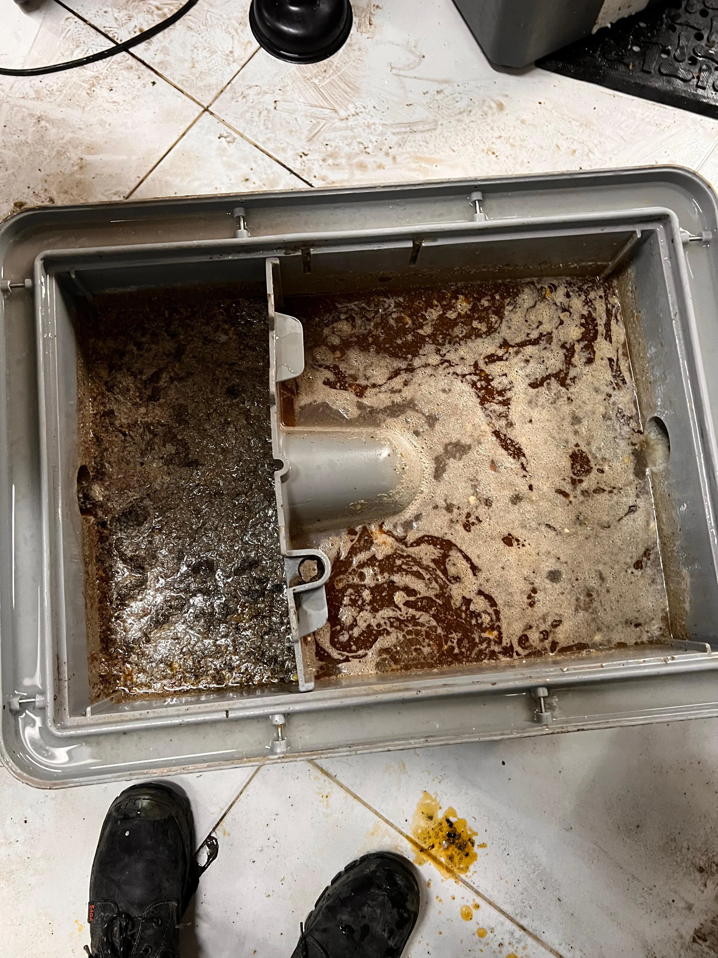 Interior de una máquina de café espresso con residuos de café y restos de mugre en una de sus secciones. Se observa también parte del suelo y zapatos de una persona.