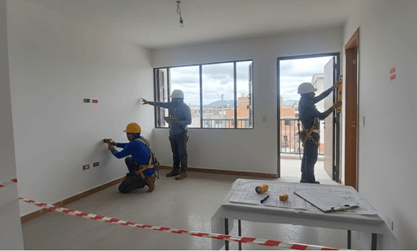 Tres trabajadores en un espacio interior en construcción, usando cascos y herramientas, realizando trabajos en las paredes y ventanas.