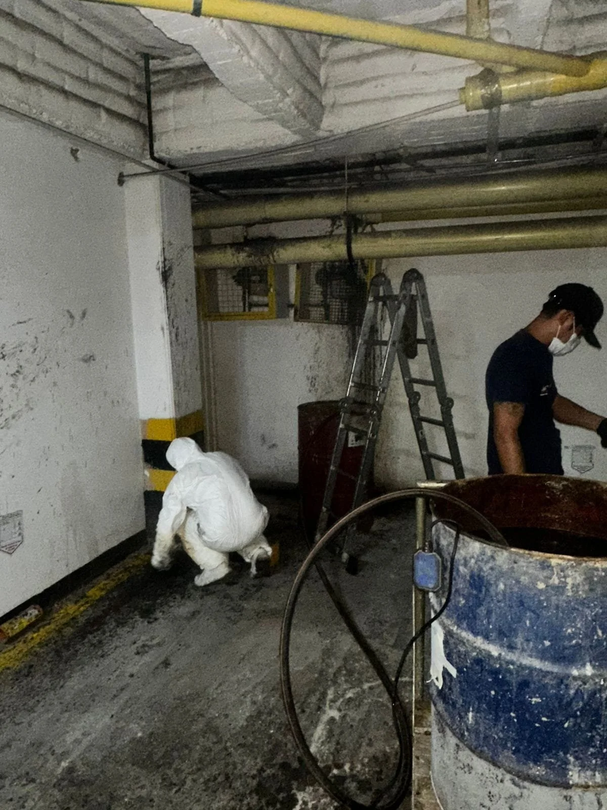 Persona con ropa de protección y mascarilla inspeccionando en un área de estacionamiento, con un perro blanco sentando en el suelo, herramientas y tuberías visibles.
