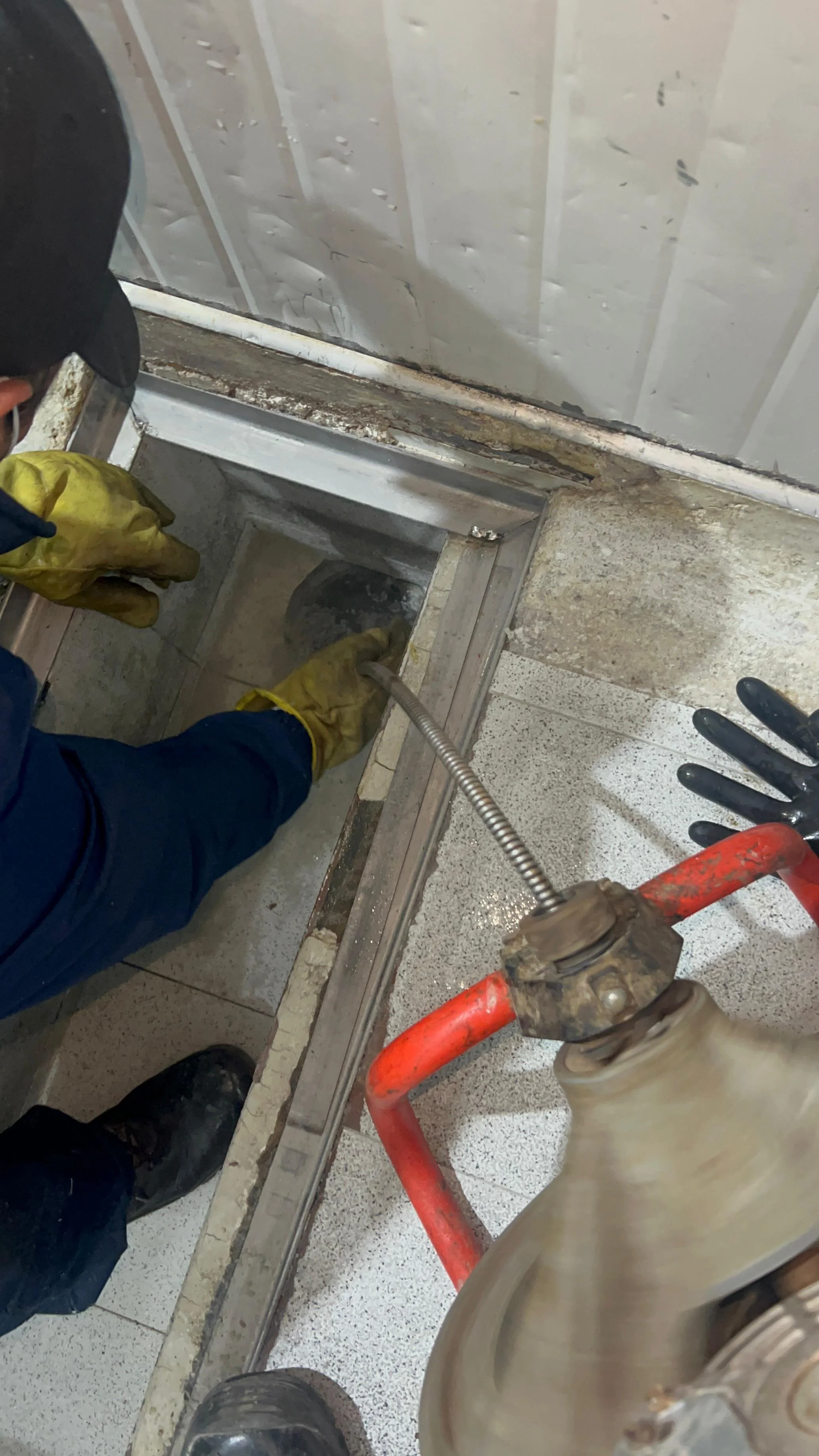 Dos personas realizan un sondeo en un acceso de piso de un edificio, una con guantes amarillos y la otra con guantes negros, usando herramientas y equipo de protección.