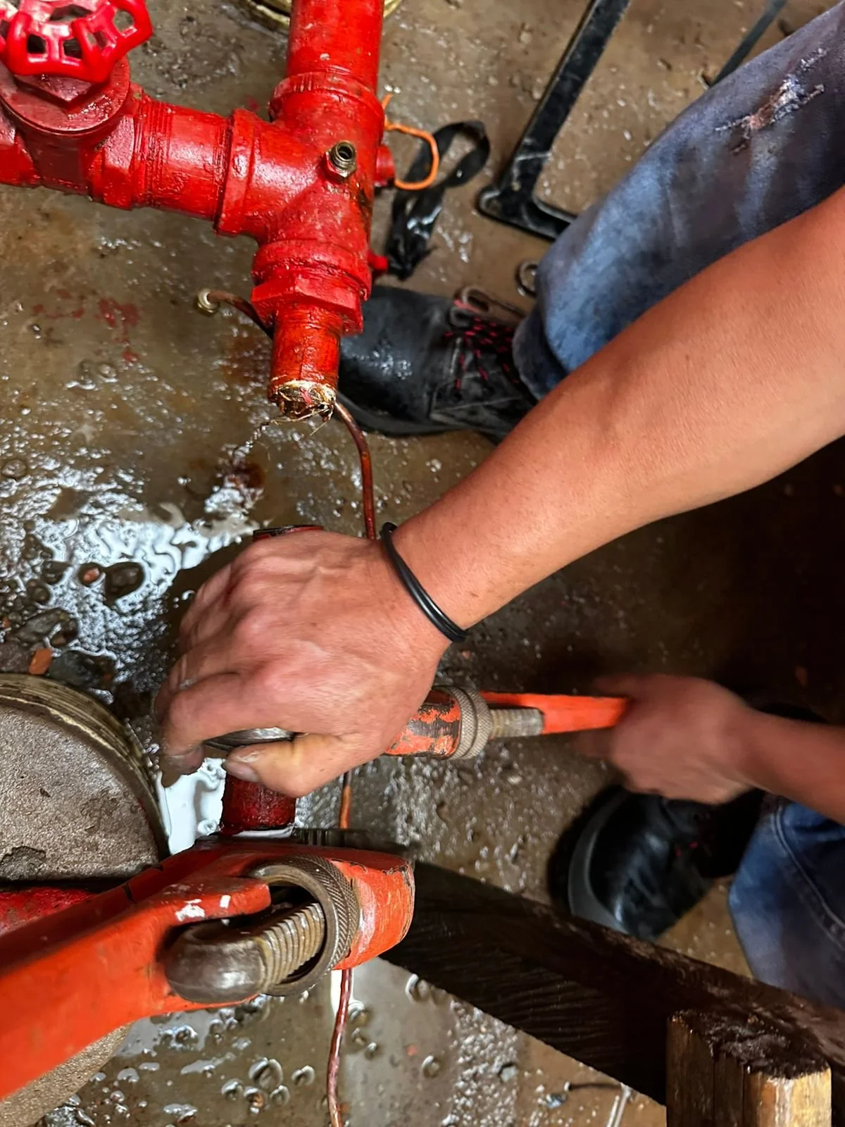 Persona reparando o manipulando una tubería de agua roja en un entorno húmedo, con herramientas y suelo mojado.