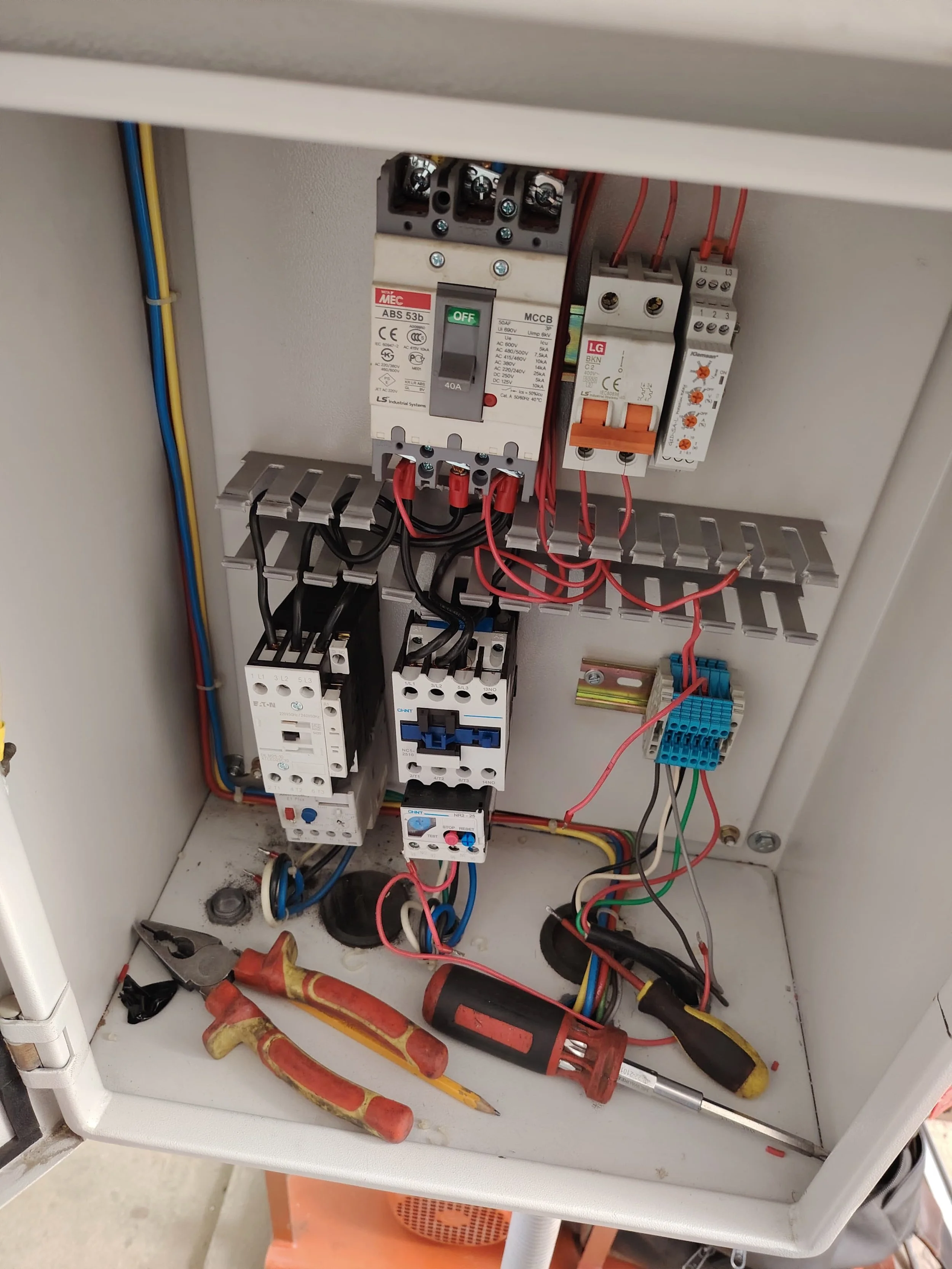 Caja de control eléctrico con componentes y herramientas dentro, incluyendo disyuntores, relés, cables de diferentes colores, y herramientas como un martillo, destornillador y pinzas en el interior.