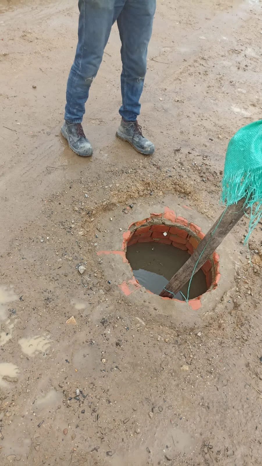 Persona con pantalones azules y botas de trabajo de pie frente a un hoyo con agua, en tierra arenosa, en una obra en construcción.