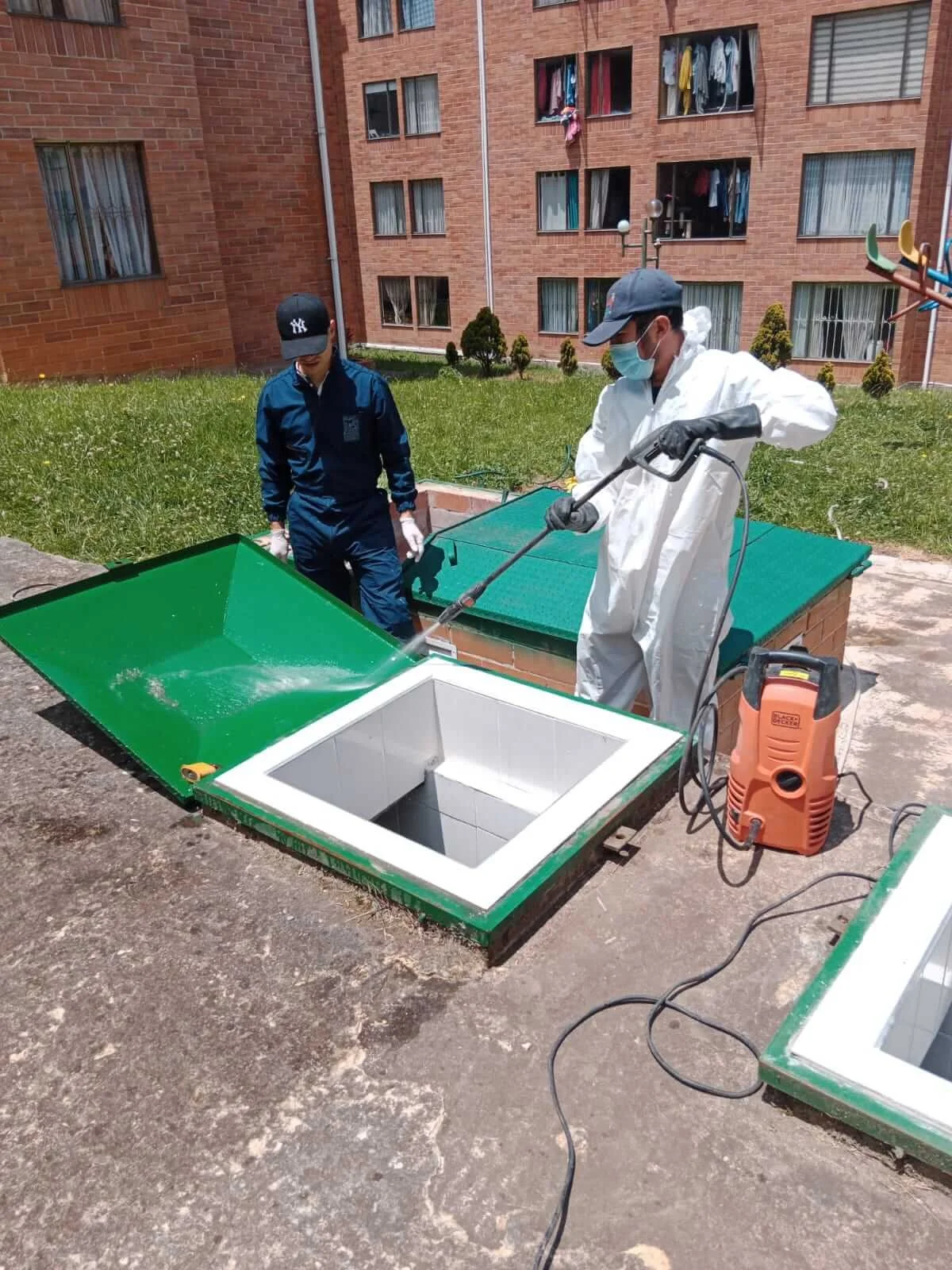 Dos personas en uniformes de protección limpiando una abertura en el suelo de un área exterior de un edificio de apartamentos.