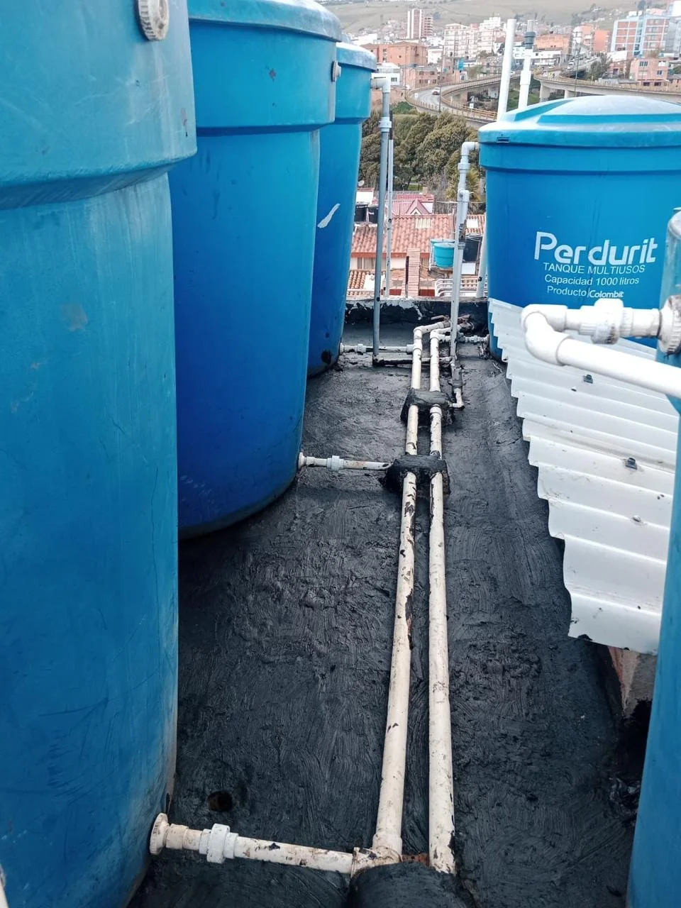 Tanques azules de agua en una azotea, con tuberías blancas conectándolos, en una zona urbana con edificios en el fondo.