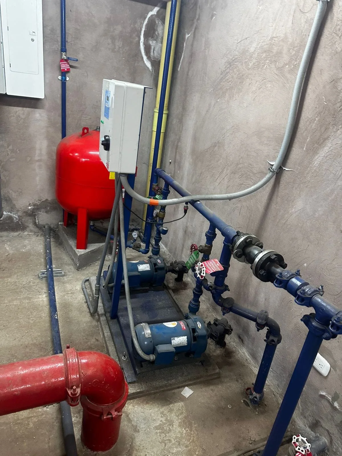 Sistema de bombeo con tubos azules y rojos, dos bombas, un tanque de presión rojo y conexiones eléctricas en una pared de cemento en un área técnica.