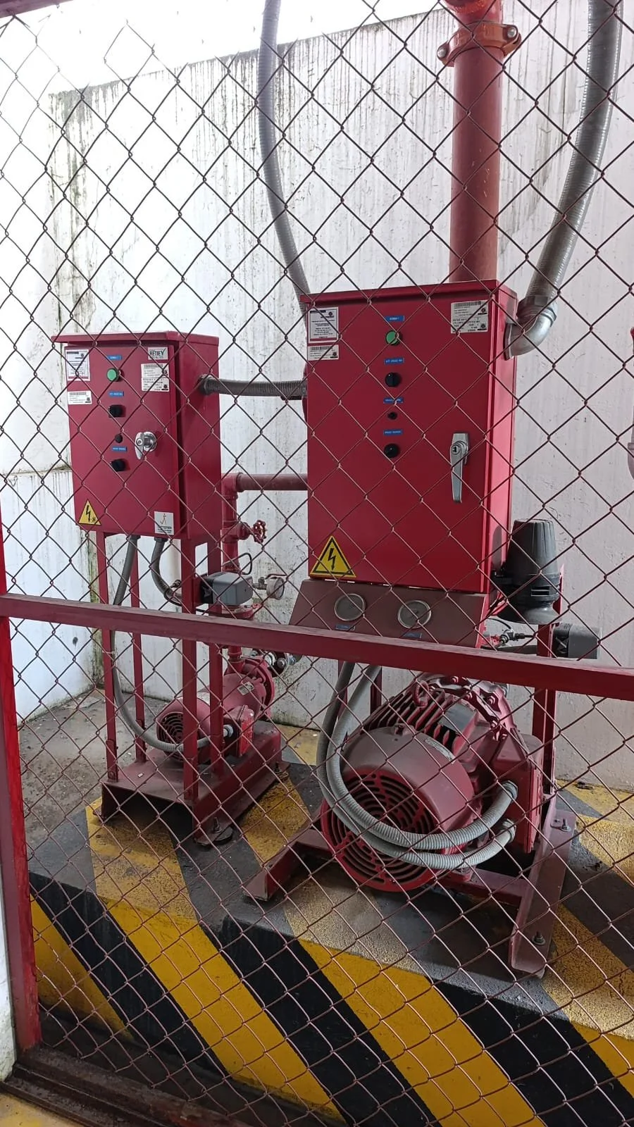 Equipo eléctrico y bombas rojas tras una cerca de malla metálica en un área industrial.