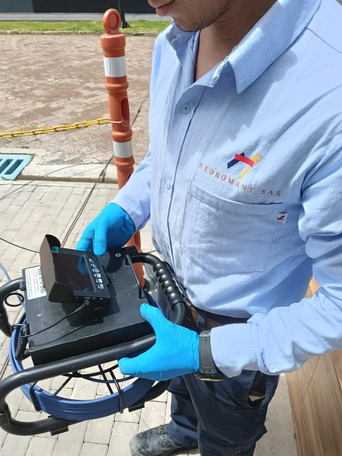Hombre con guantes azules operando un equipo en la calle, con un chaleco que dice 'Hydromant SAS' Realizando un servicio de geófono