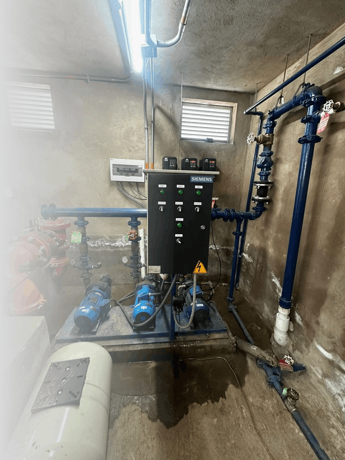 Sistema de bombeo con bombas y tuberías de agua en una habitación de cemento con ventanas pequeñas