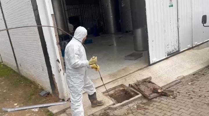 Persona con traje protector y guantes inspecciona y abre una tapa de un alcantarillado en la acera frente a un almacén.