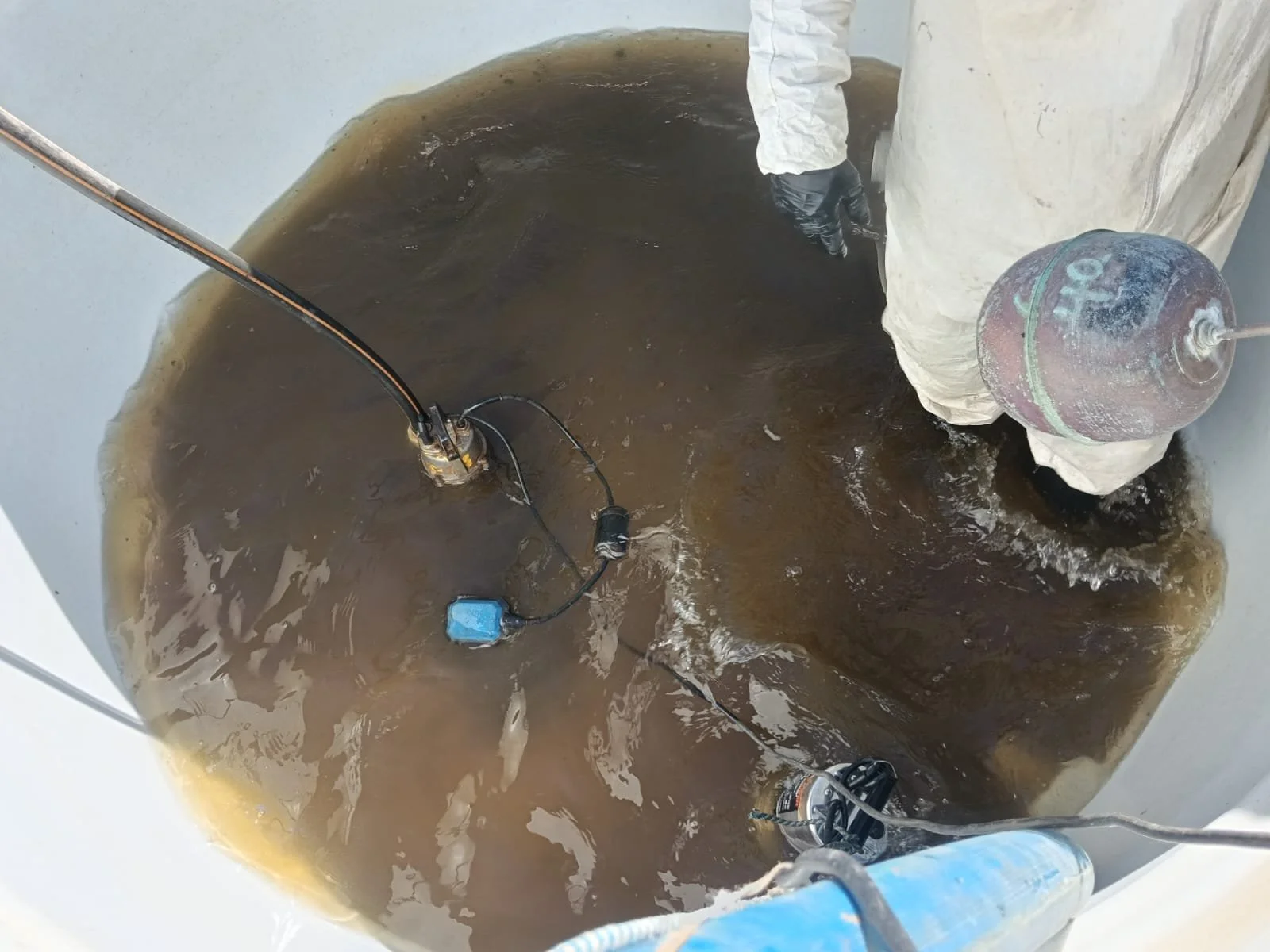 Una persona en traje protector y guantes, sumergida en un tanque con agua oscura, parece estar operando una bomba sumergible para limpiar o drenar el tanque.
