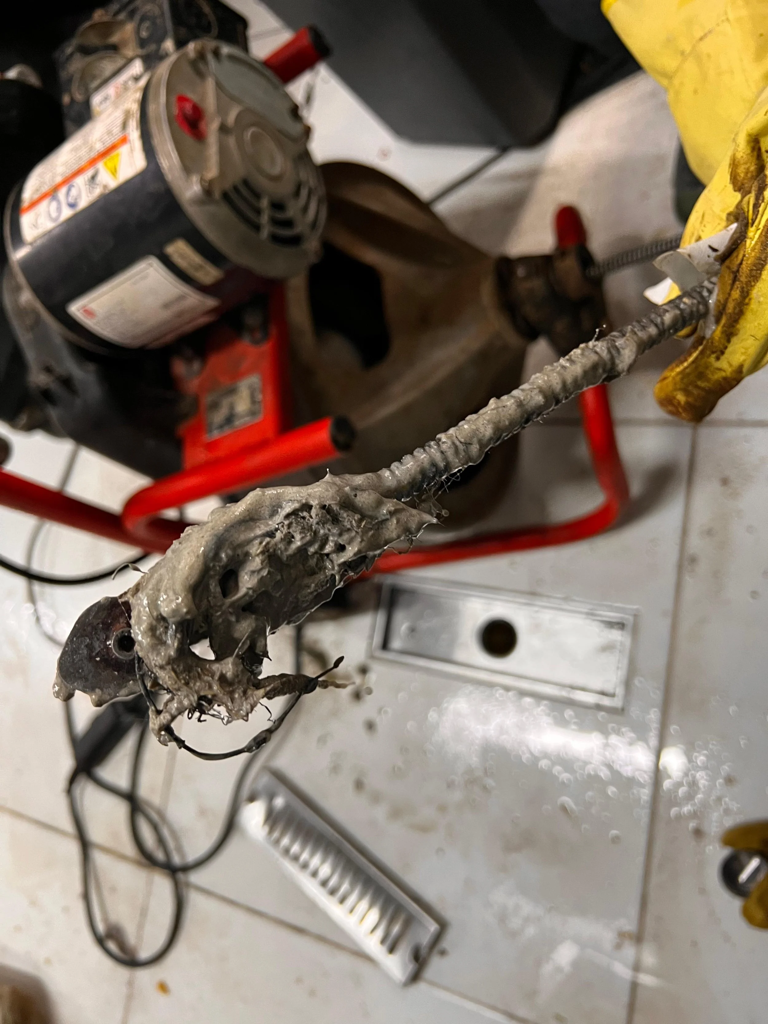 Un objecto metálico con apariencia quemada y deformada, sostenido con pinzas amarillas, en un taller con suelo de baldosas y herramientas cercanas.