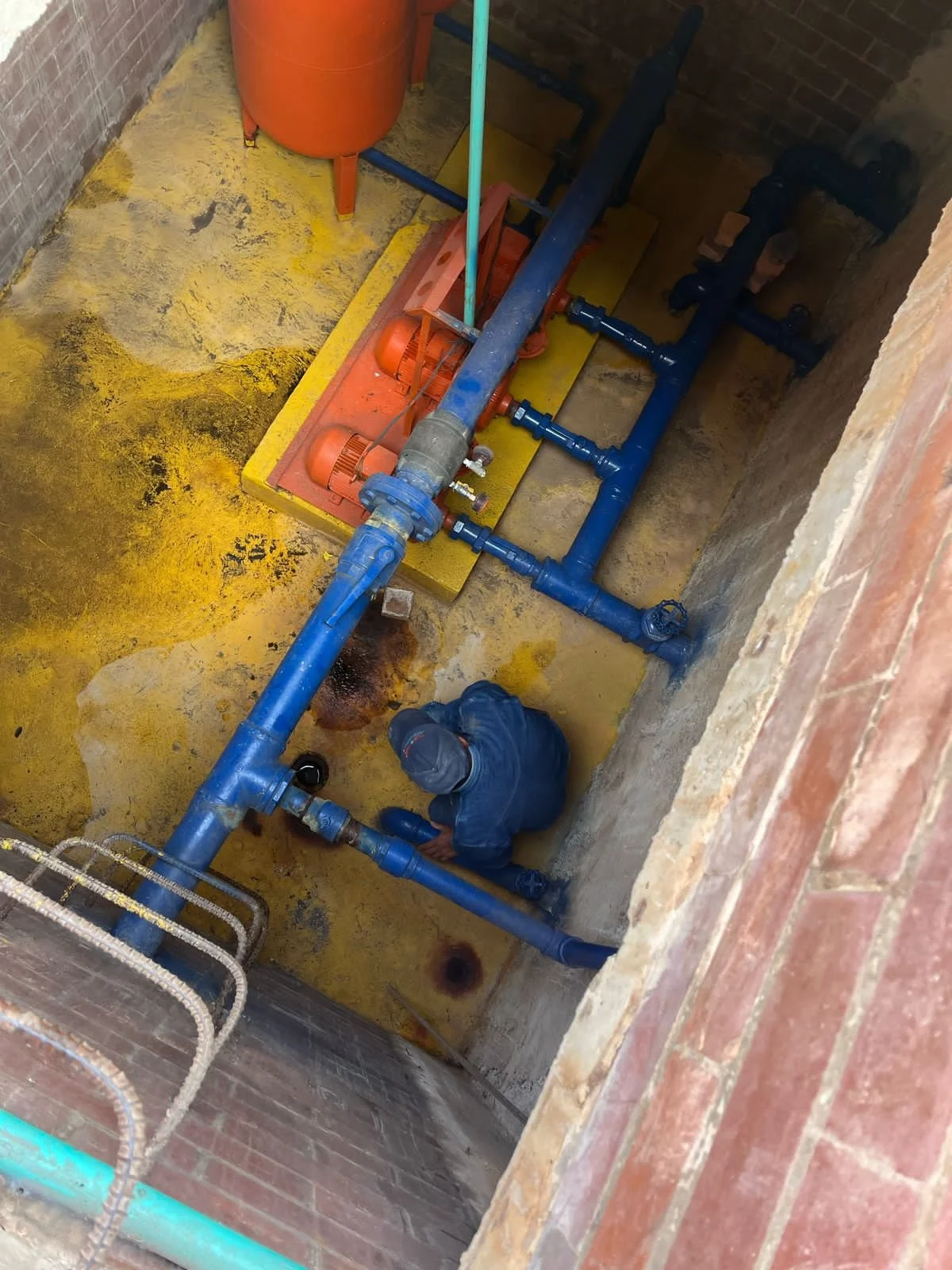 Técnico realizando mantenimiento en un sistema de tuberías de agua o gas en una construcción, en un área de pozo con paredes de ladrillo y piso color amarillo