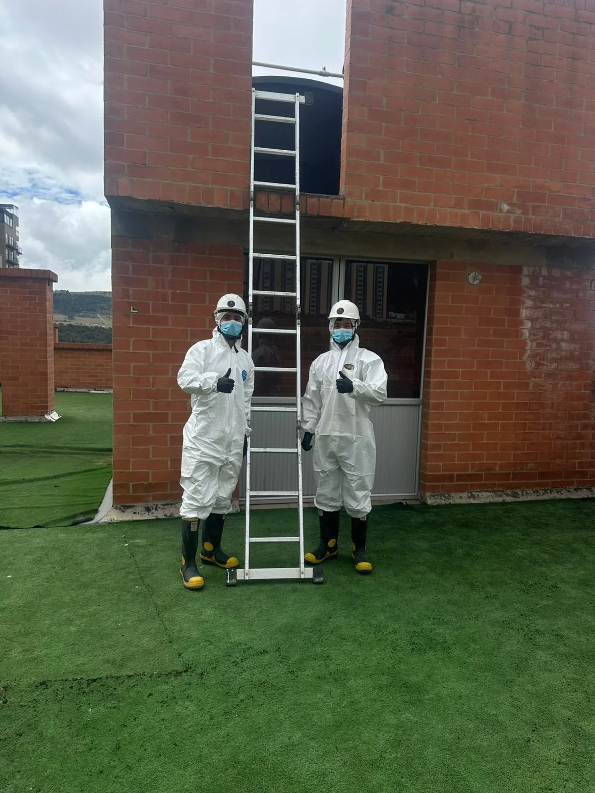Dos trabajadores con trajes de protección, guantes, máscaras y botas de seguridad, de pie en un techo con césped artificial, sosteniendo una escalera y haciendo señal de pulgar hacia arriba para lavar tanques de agua potable
