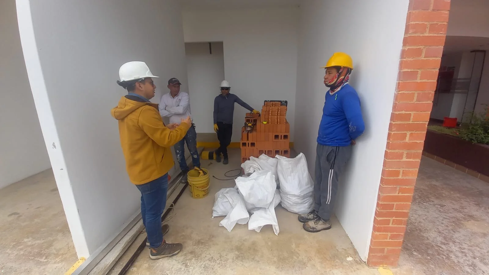Cuatro trabajadores de construcción con cascos y ropa de trabajo discuten en una obra, con ladrillos, bolsas y herramientas a su alrededor.