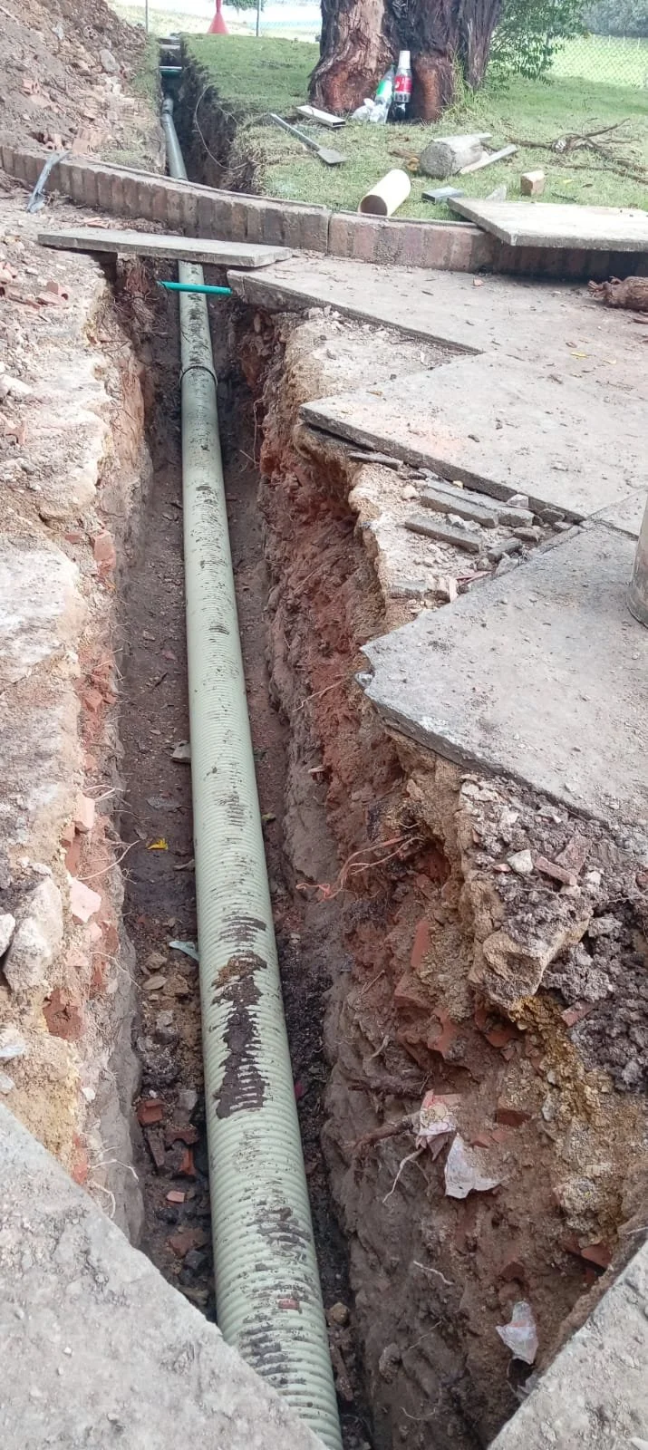 Tubo de alcantarillado instalado en una zanja en proceso de construcción con tierra y ladrillos a los lados, rodeado de cemento y herramientas de construcción.