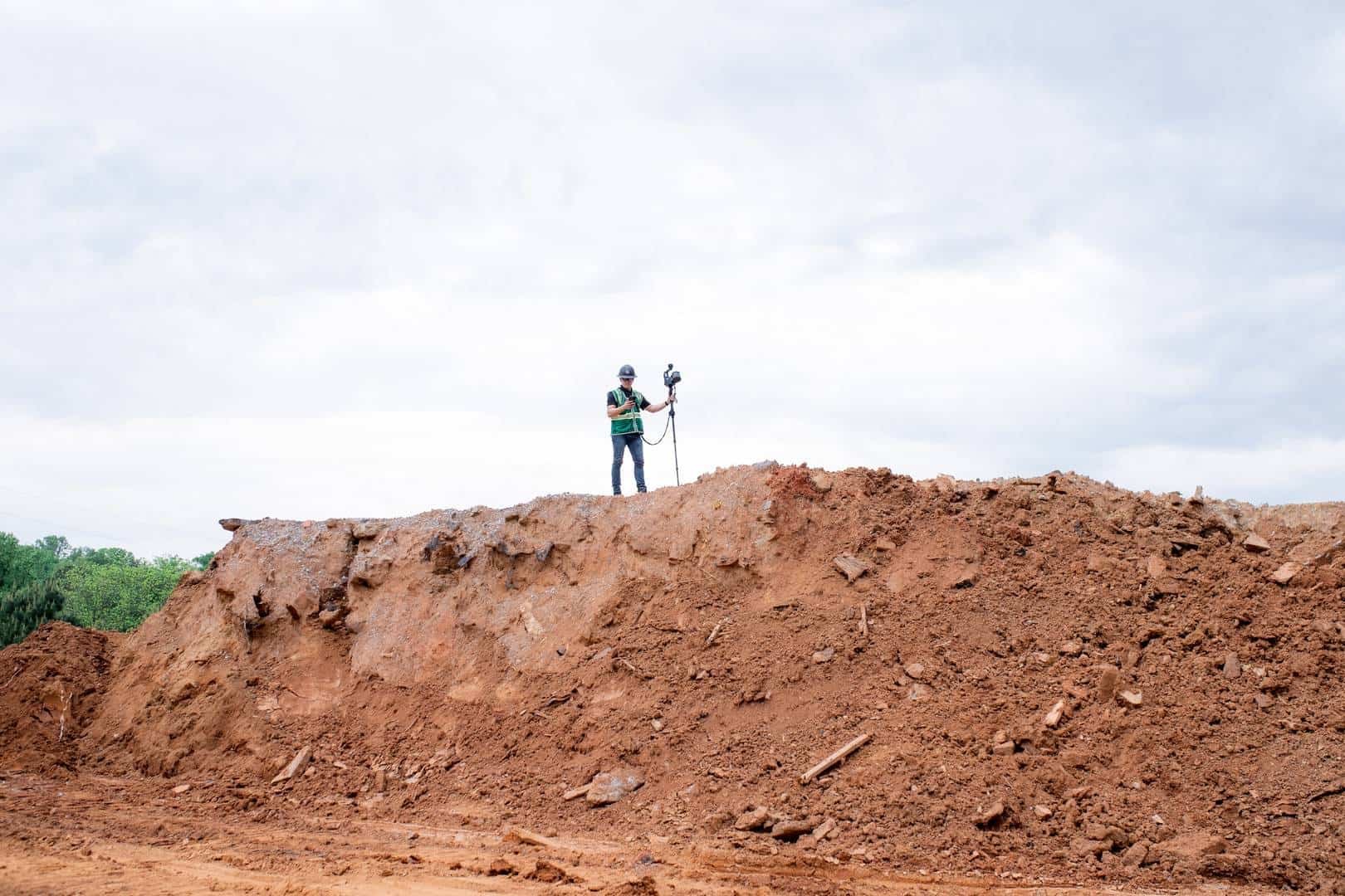 Un hombre investido con casco y chaleco de seguridad sosteniendo equipo de medición sobre un terreno de tierra en un área de construcción o exploración del suelo