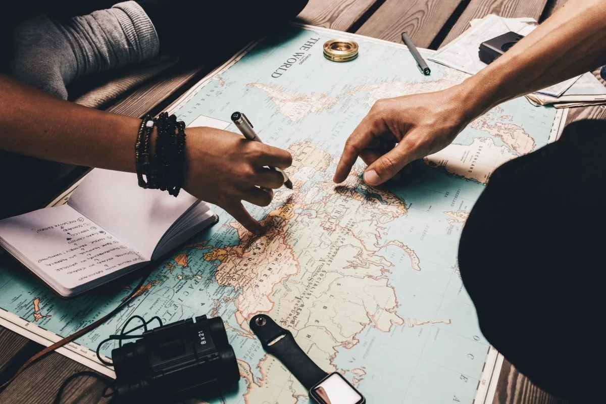 Two people planning a trip on a world map, with one person pointing to a location and the other writing notes.