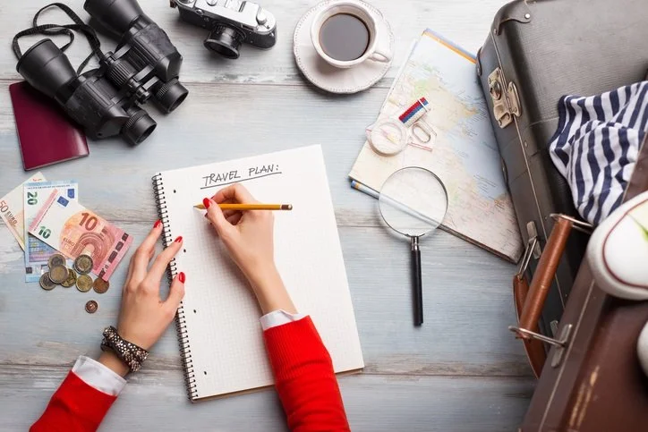 Overhead view of a travel planning scene with a person's hands writing in a notebook labeled 'Travel Plan', surrounded by binoculars, camera, cup of coffee, map, magnifying glass, summarization of money and coins, and a partially packed suitcase with clothes.