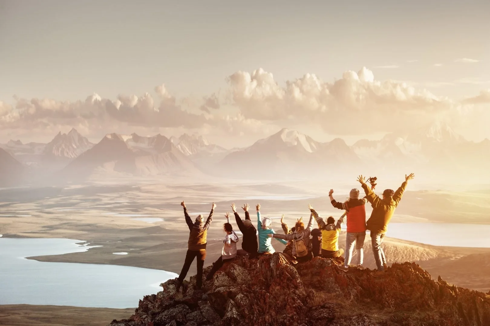 Group of people celebrating on top of a mountain ridge with mountains and lakes in the background at sunrise or sunset.