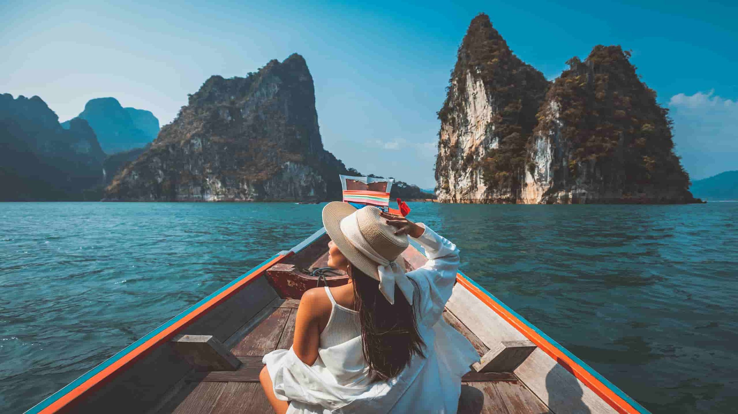 A woman in a white dress and wide-brimmed hat sitting on a boat, gazing at limestone islands in a body of water.