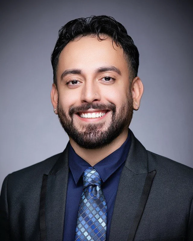 Portrait of a smiling man with a beard, dark hair, earrings, wearing a dark suit, blue dress shirt, and patterned blue tie, against a gray background.