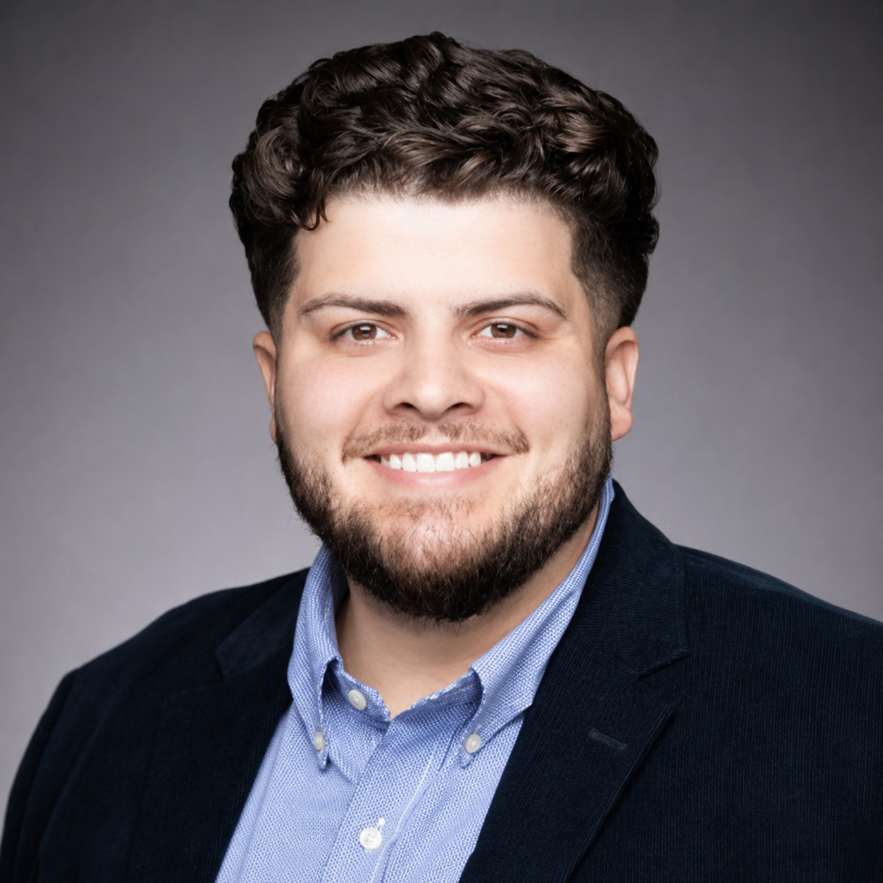 Portrait of a man with dark, curly hair, a beard, and light skin, wearing a dark blazer and a light blue button-up shirt smiling against a gray background.