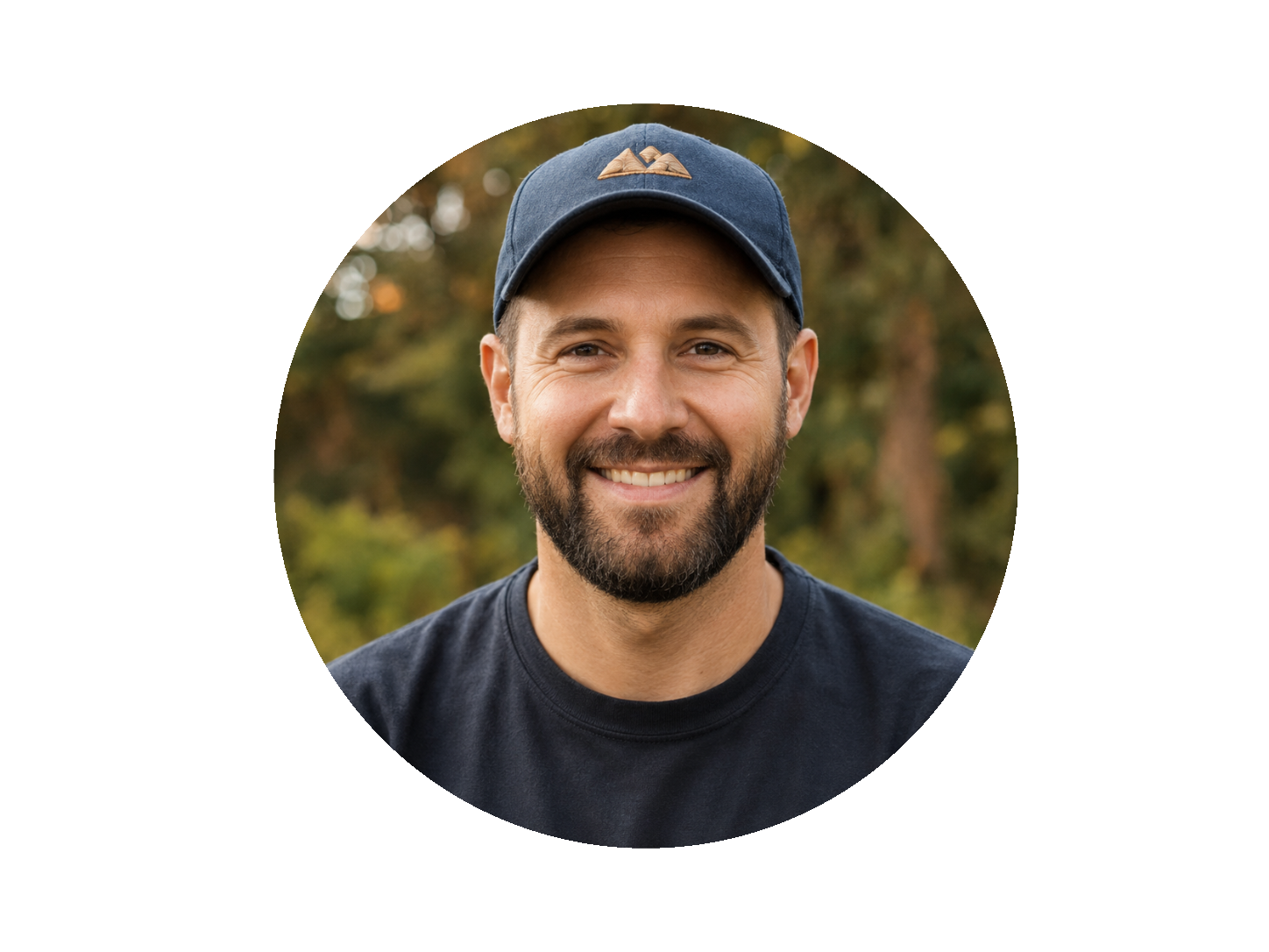 A smiling man with a beard, wearing a navy blue baseball cap and a black t-shirt, outdoors with trees in the background.