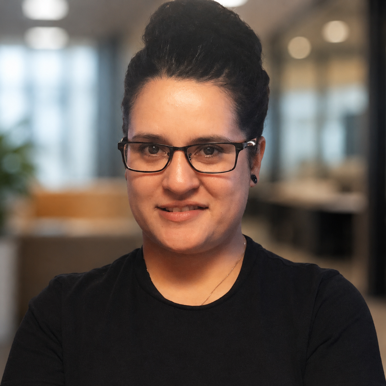 A woman with dark hair in a bun, wearing glasses, a black shirt, and small earrings, smiling in an indoor setting with blurred background.