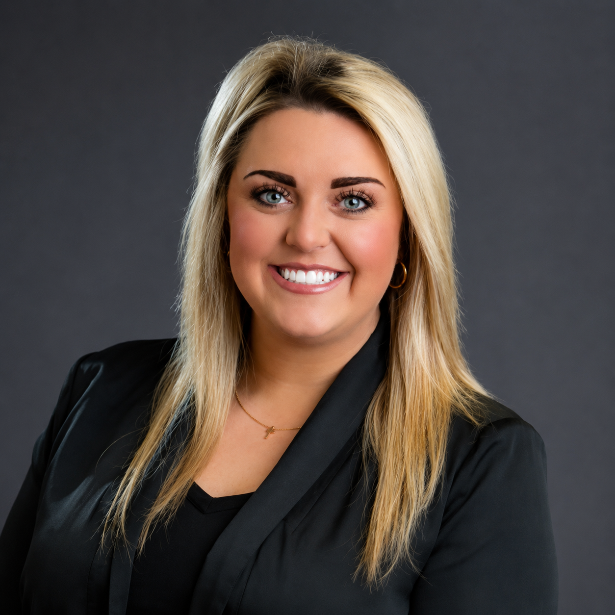 A professional woman with long blonde hair, blue eyes, and fair skin, smiling, wearing a black blazer, a gold necklace, and gold hoop earrings, against a dark gray background.