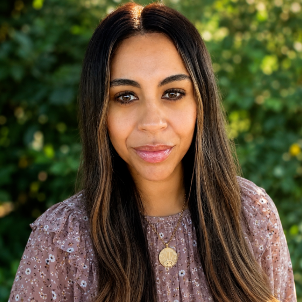 Close-up portrait of a young woman with long dark hair and brown eyes, wearing a floral blouse and layered gold necklaces, outdoors with green foliage in the background.