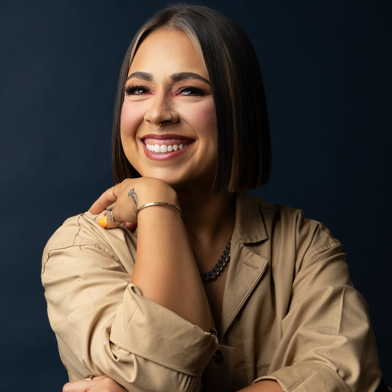 A young woman with shoulder-length dark hair, wearing a beige shirt, smiling with her teeth showing, resting her chin on her hand. She has a nose ring, jewelry on her fingers and wrist, and a tattoo on her wrist.