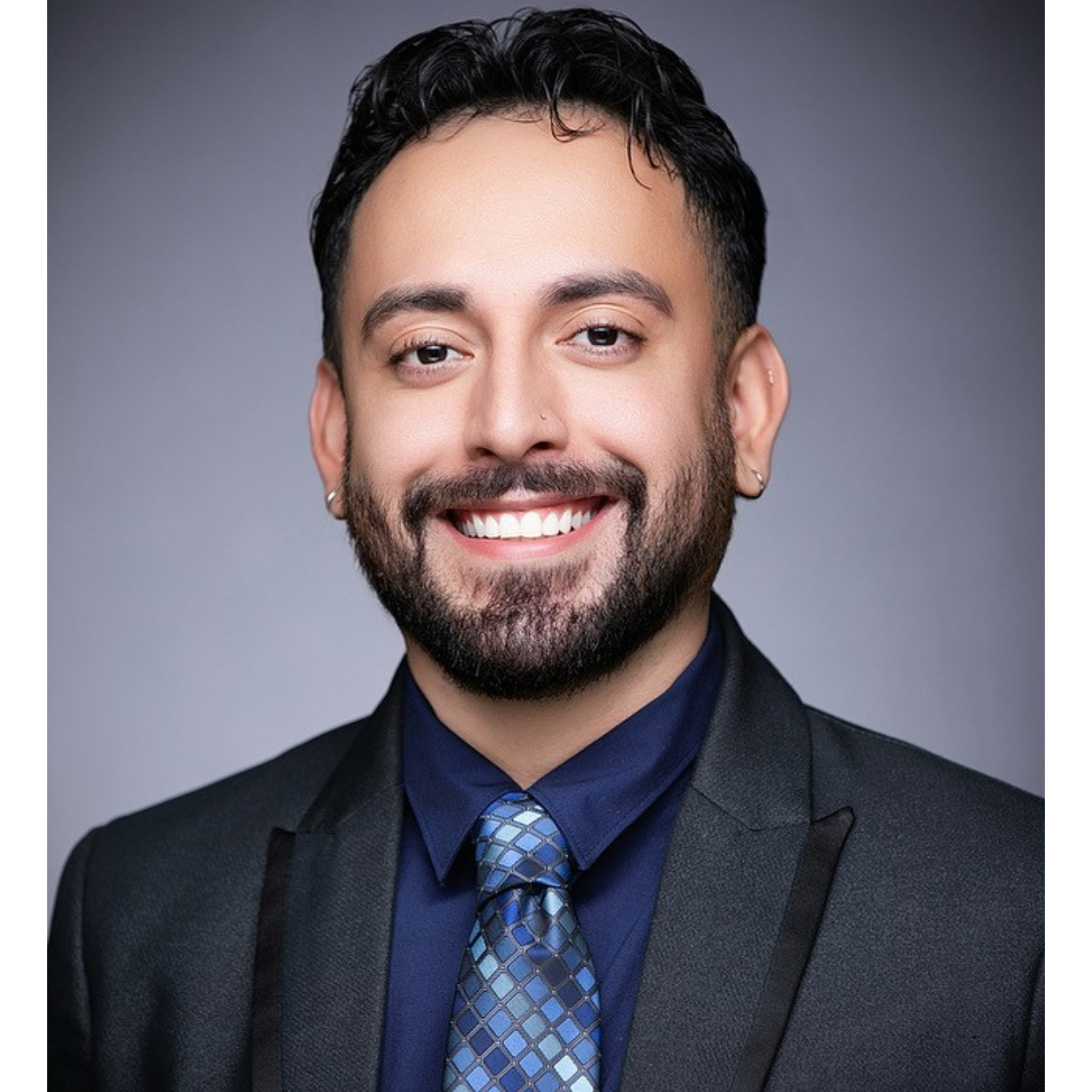 A smiling man with dark hair and a beard, wearing a dark suit, blue shirt, and patterned tie, against a gray background.