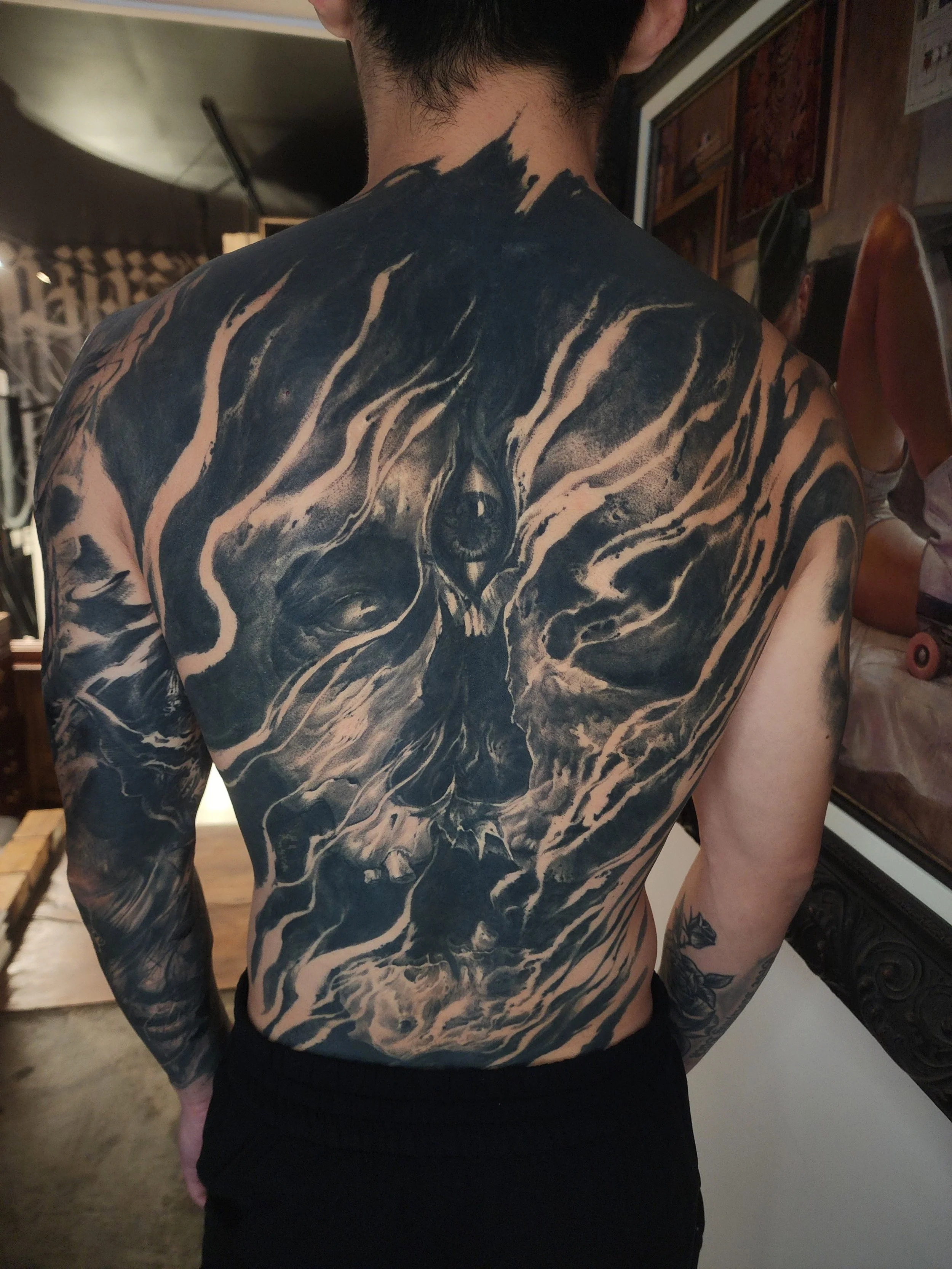 A person with a detailed black and white tiger tattoo covering the back, shoulders, and upper arms, standing indoors with various decorations and objects in the background.