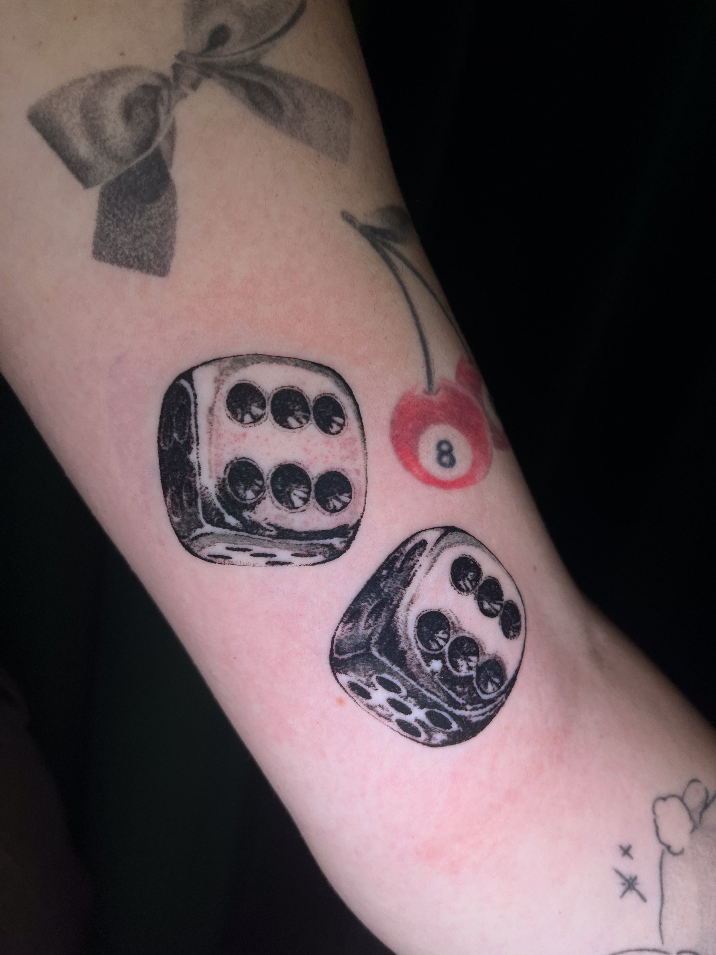 Tattoo of three black dice with white dots, and a red cherry with the number 8 on it, on a person's arm.