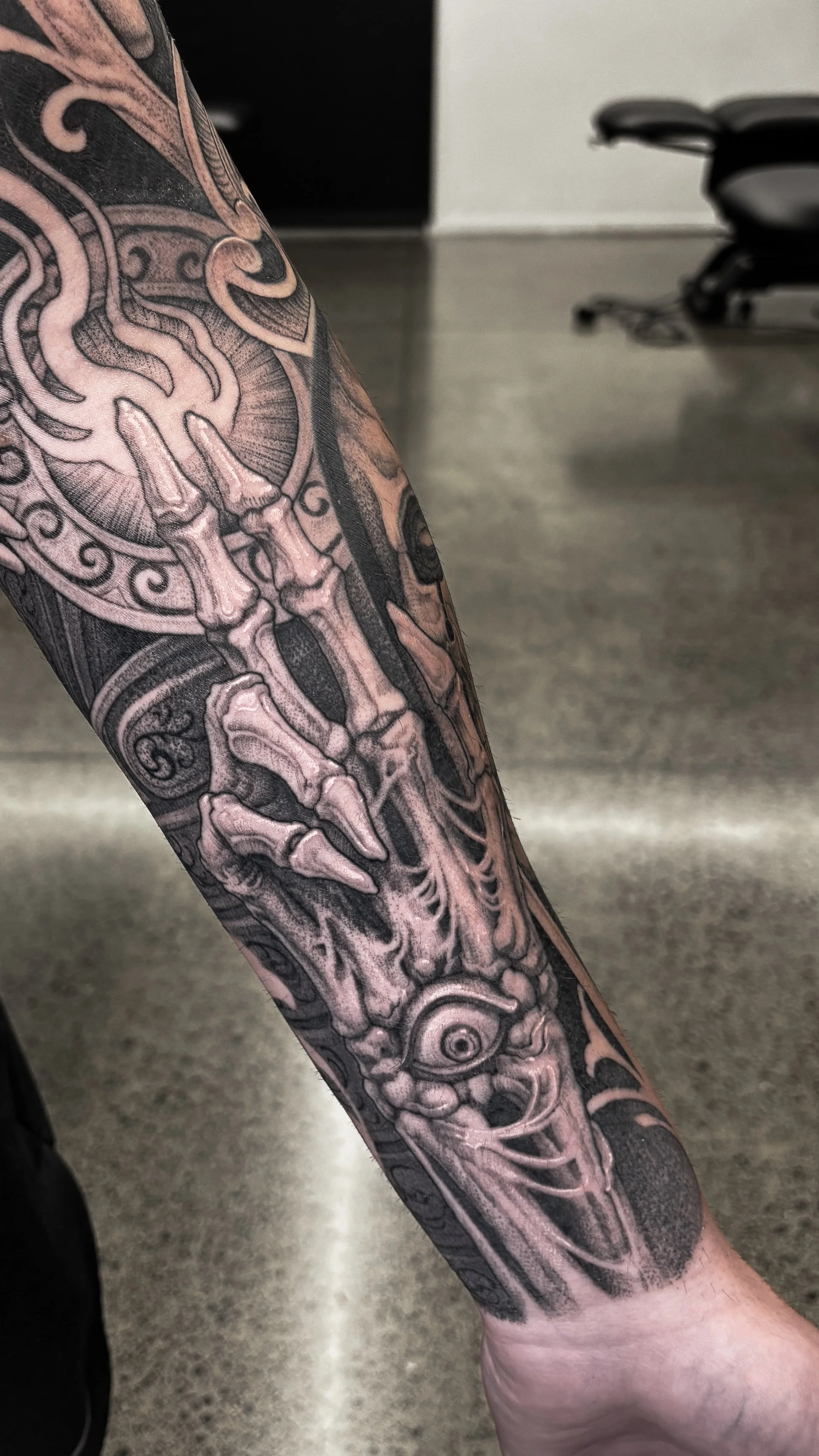Detailed black and gray tattoo on a forearm featuring a skeleton hand gripping an antique clock, with intricate shading and design elements.
