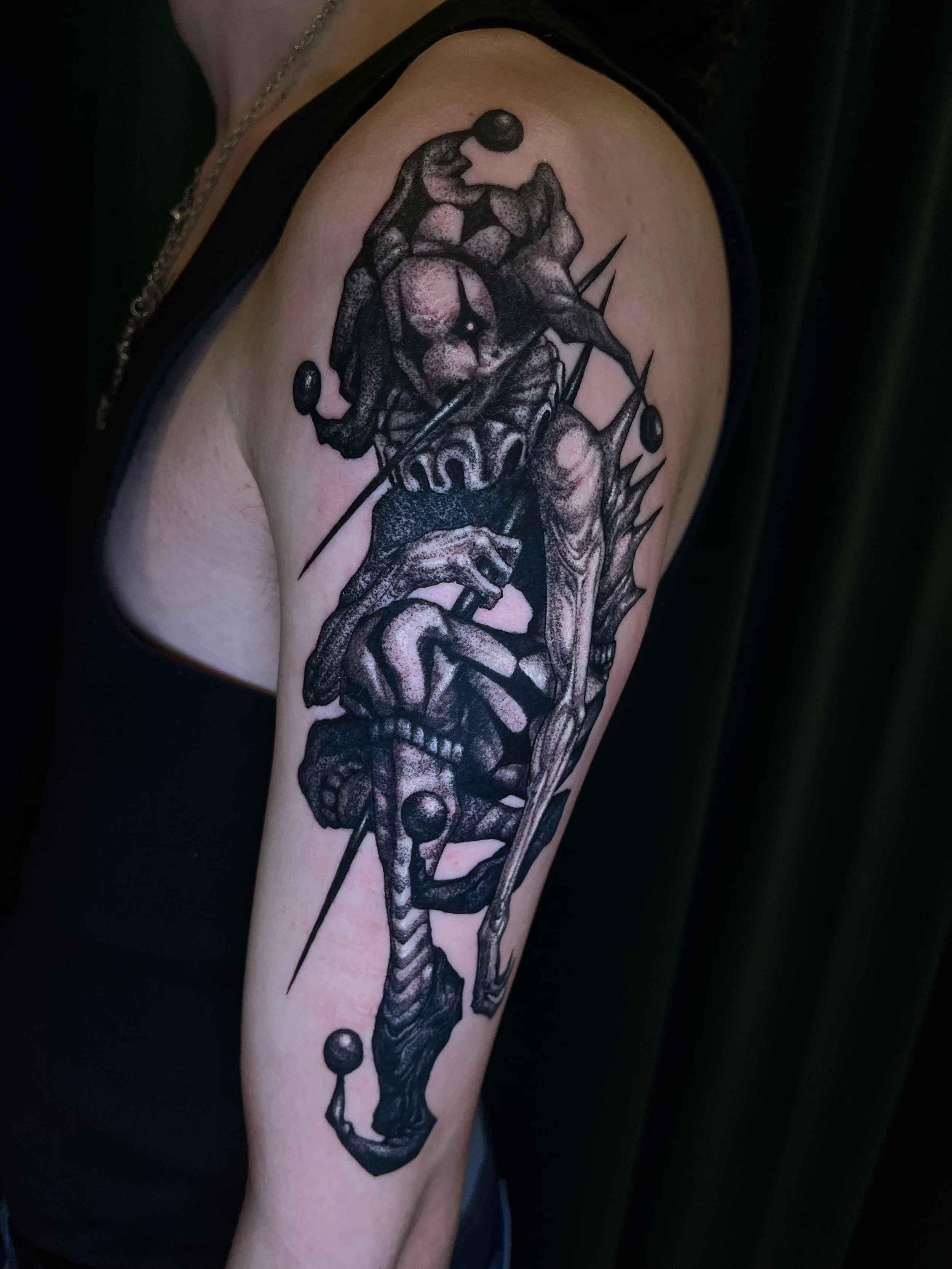 A black tattoo on a person's upper arm depicting a skeleton with skeletal hands holding a sword, a skull, and various abstract elements.