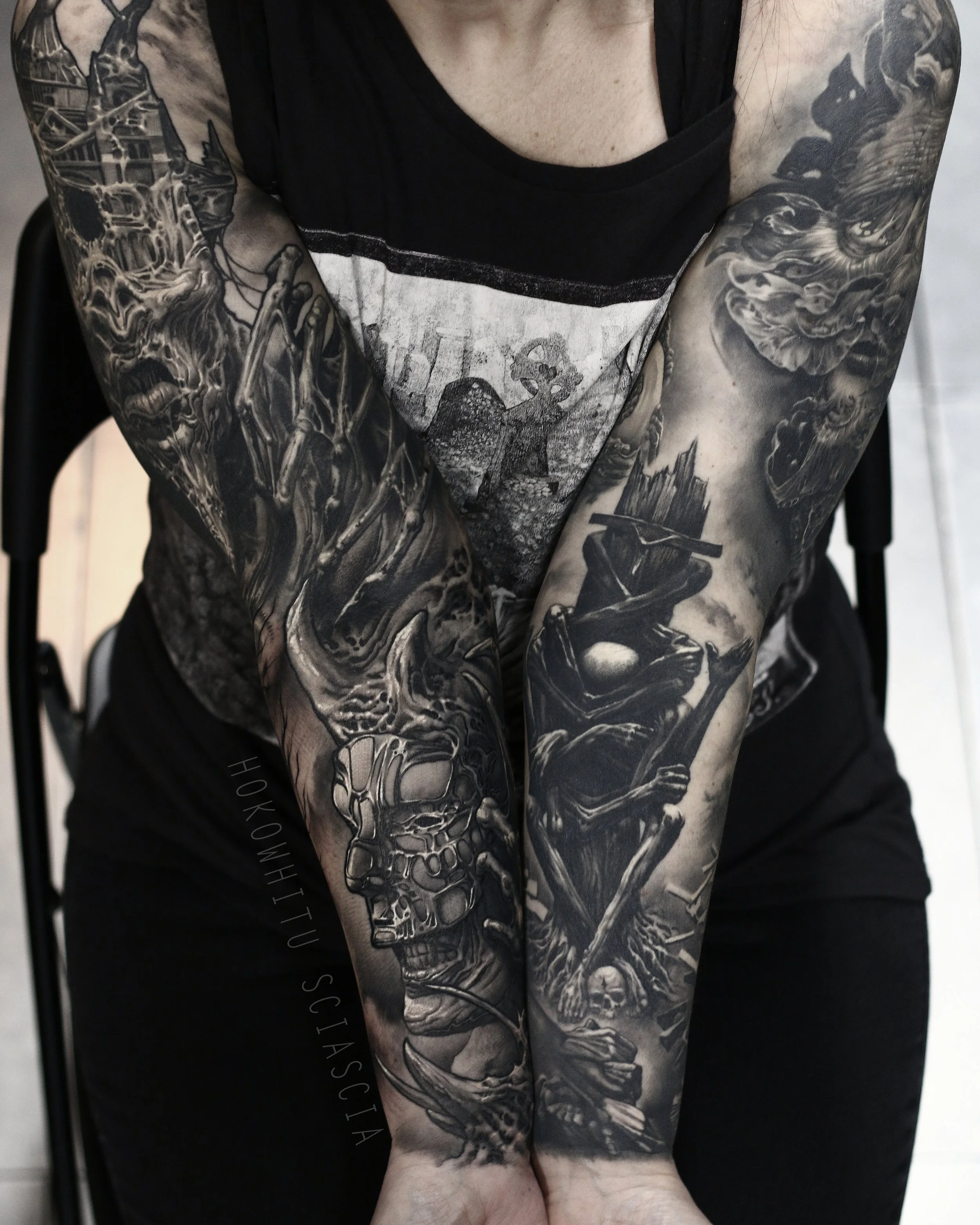 Close-up of a person's tattooed arms featuring dark, detailed black and gray horror-themed artwork, including a skull, a skeleton, and other macabre elements.