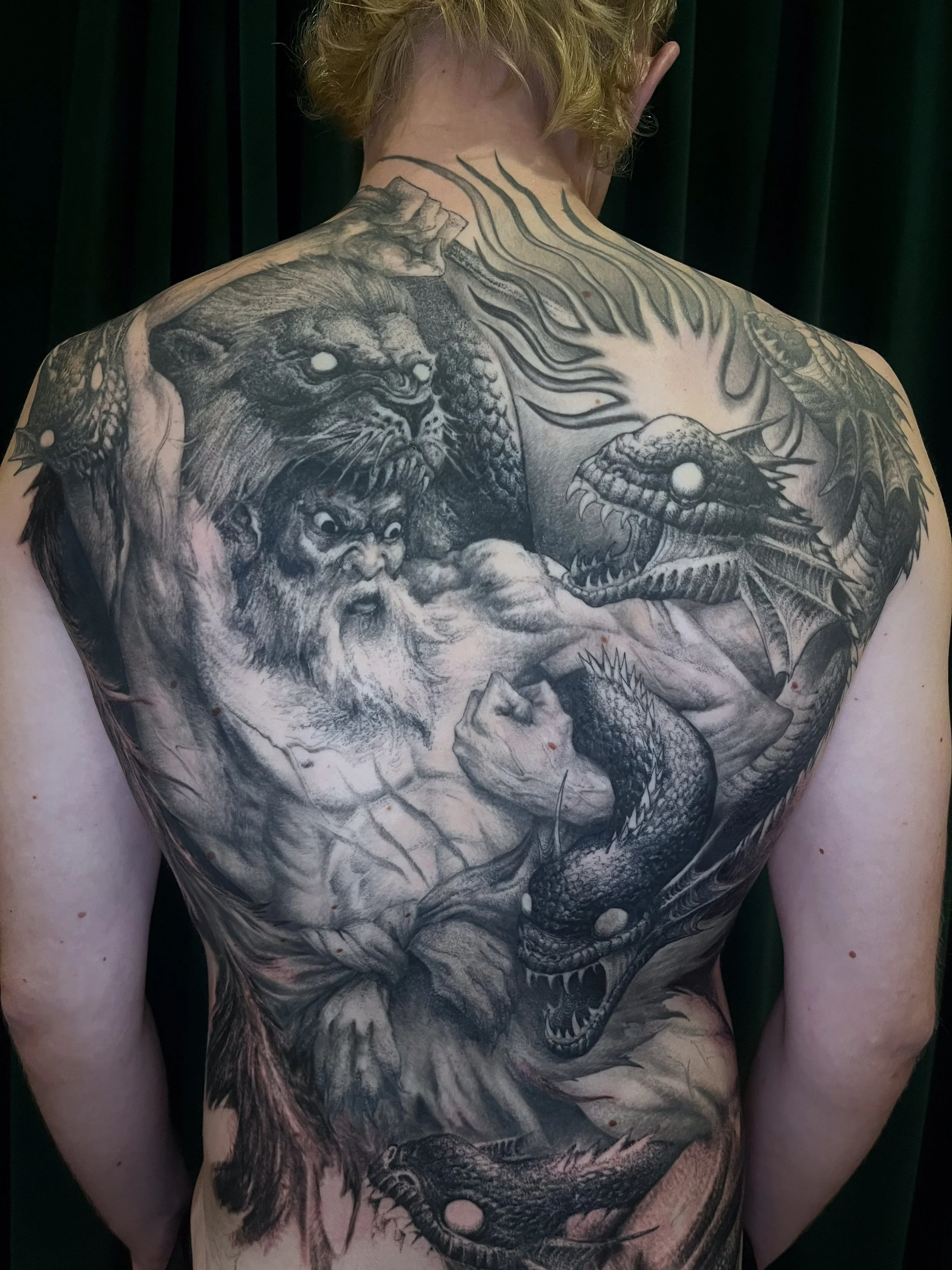 A person's back with a large black and gray tattoo of a dragon, a lion, a serpent, and other mythical creatures and figures, with detailed shading and linework.