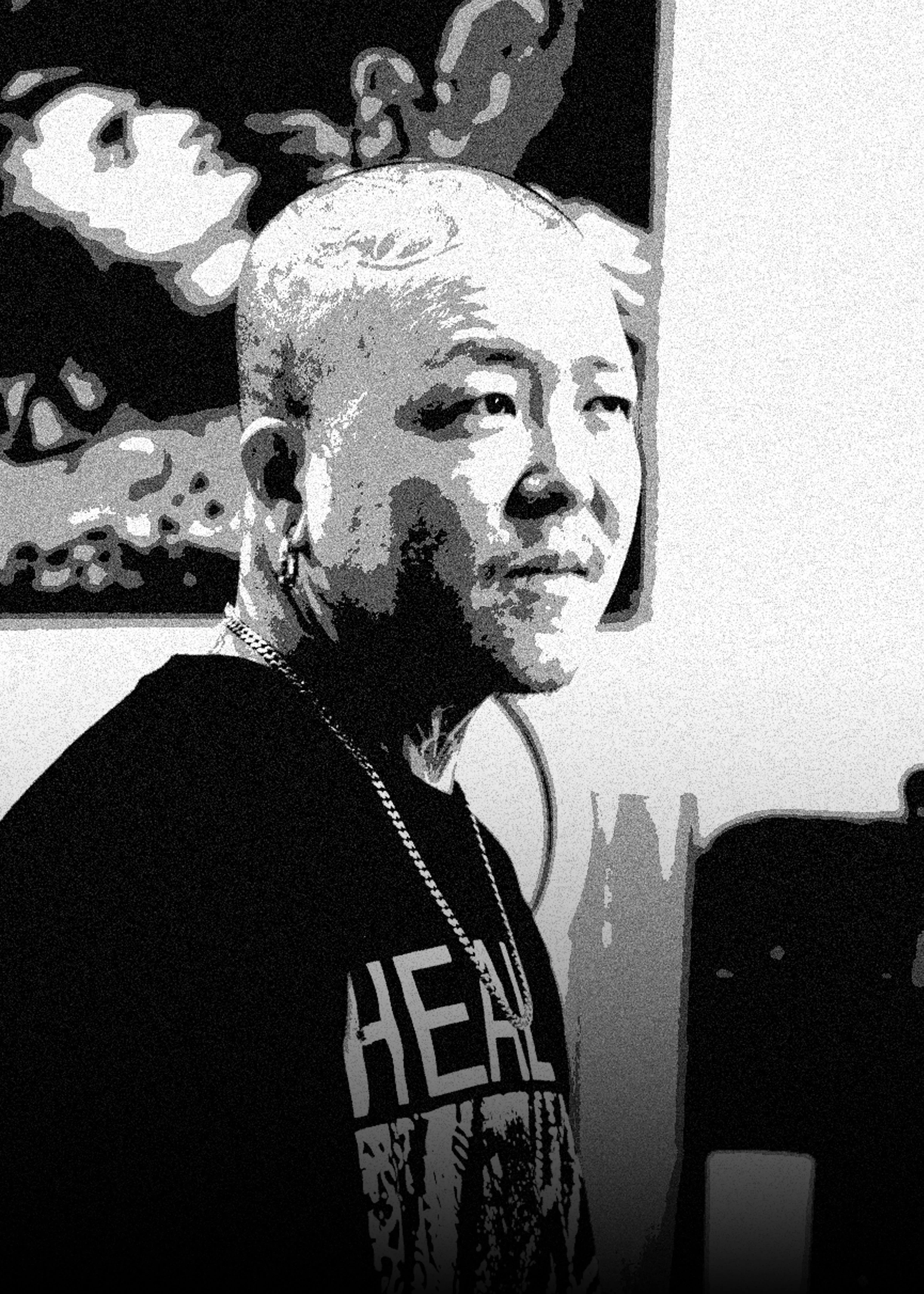 A grayscale photo of a woman with short, light-colored hair, wearing earrings, a necklace, and a hoodie with the word "HEART" visible on it. She has her eyes closed or looking downward and is in front of a background featuring a framed painting or ph