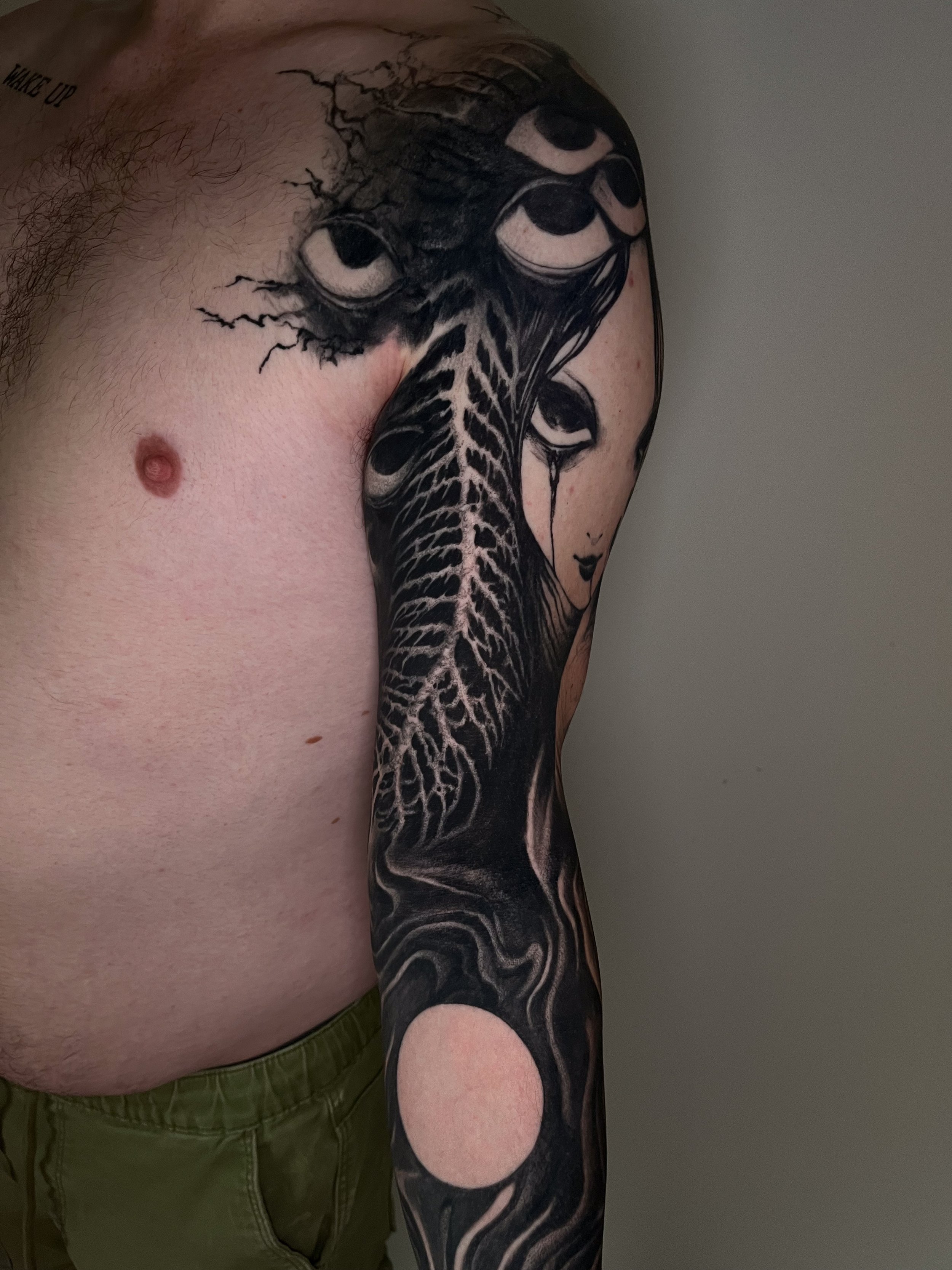 A person with a black and gray tattoo sleeve on their arm, featuring abstract eye designs, and a tattoo on the neck that IDs as 'WAKE UP'.