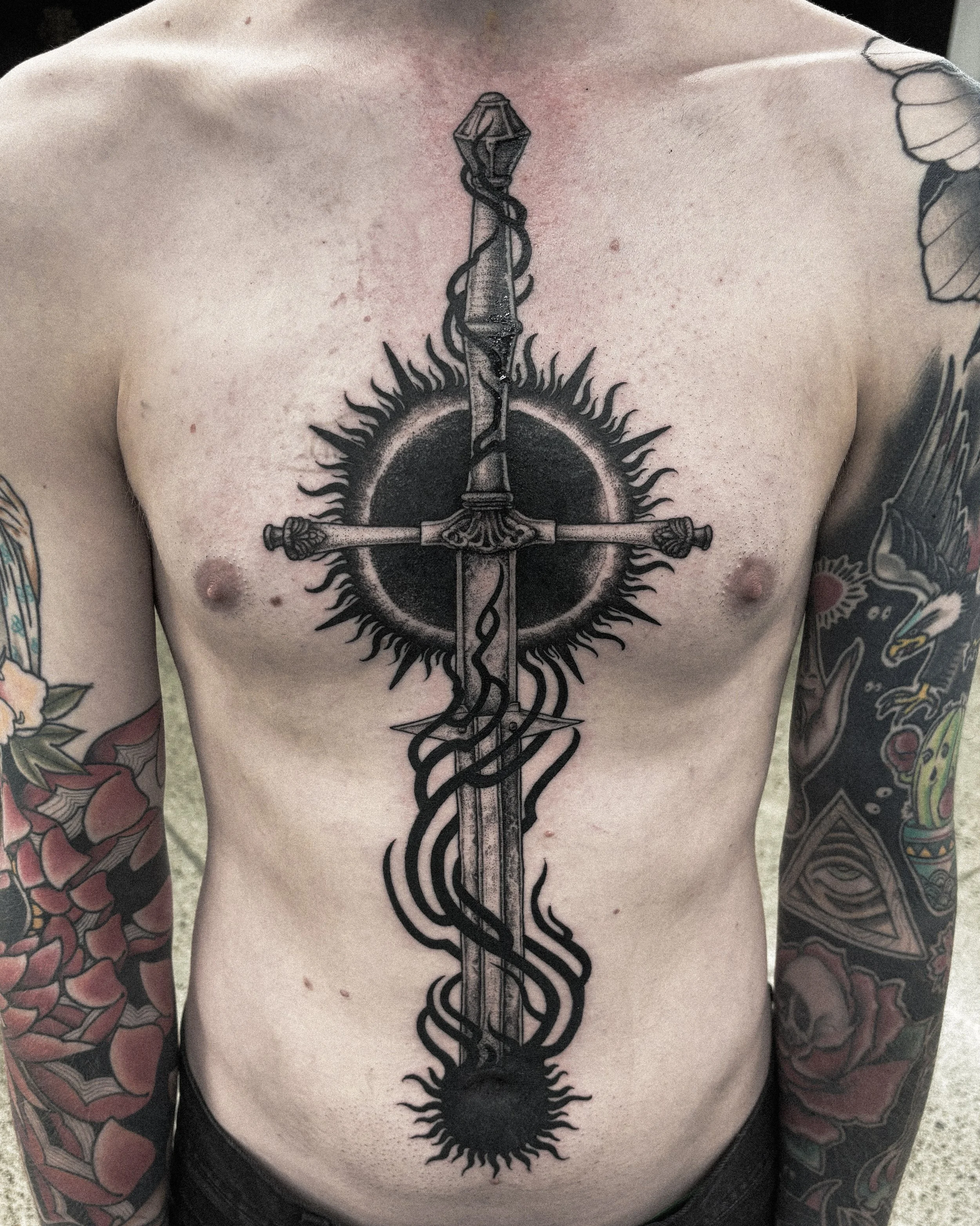 Tattoo of a sword with a decorated hilt and a snake wrapped around the blade, intersecting a circular element with spikes, located on a man's chest.