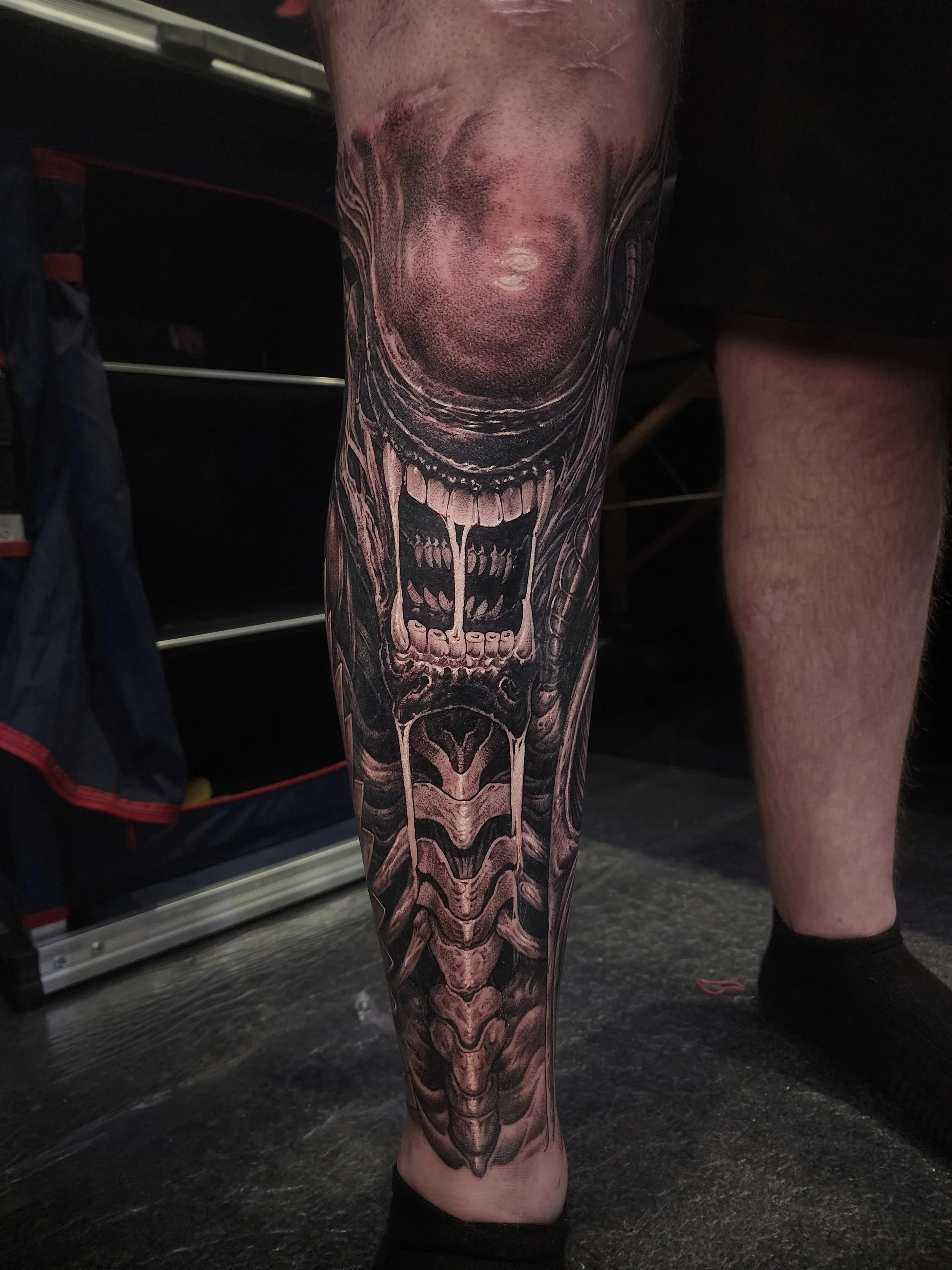 A detailed black and gray tattoo of a terrifying alien or creature with a large open mouth, sharp teeth, and skeletal features on a person's leg.