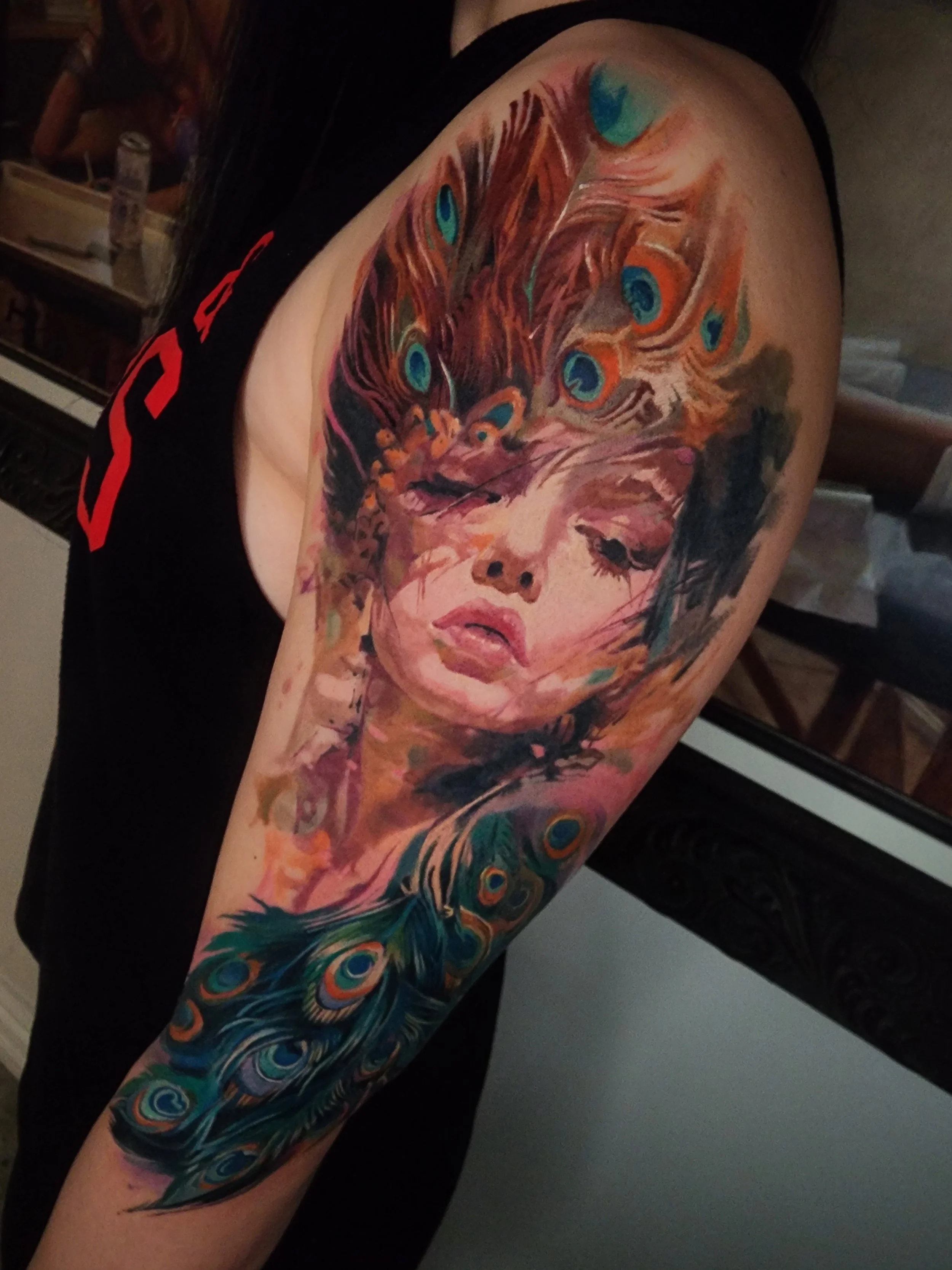 Colorful tattoo of a woman's face with closed eyes and long dark hair, surrounded by peacock feathers and peacock eye patterns.