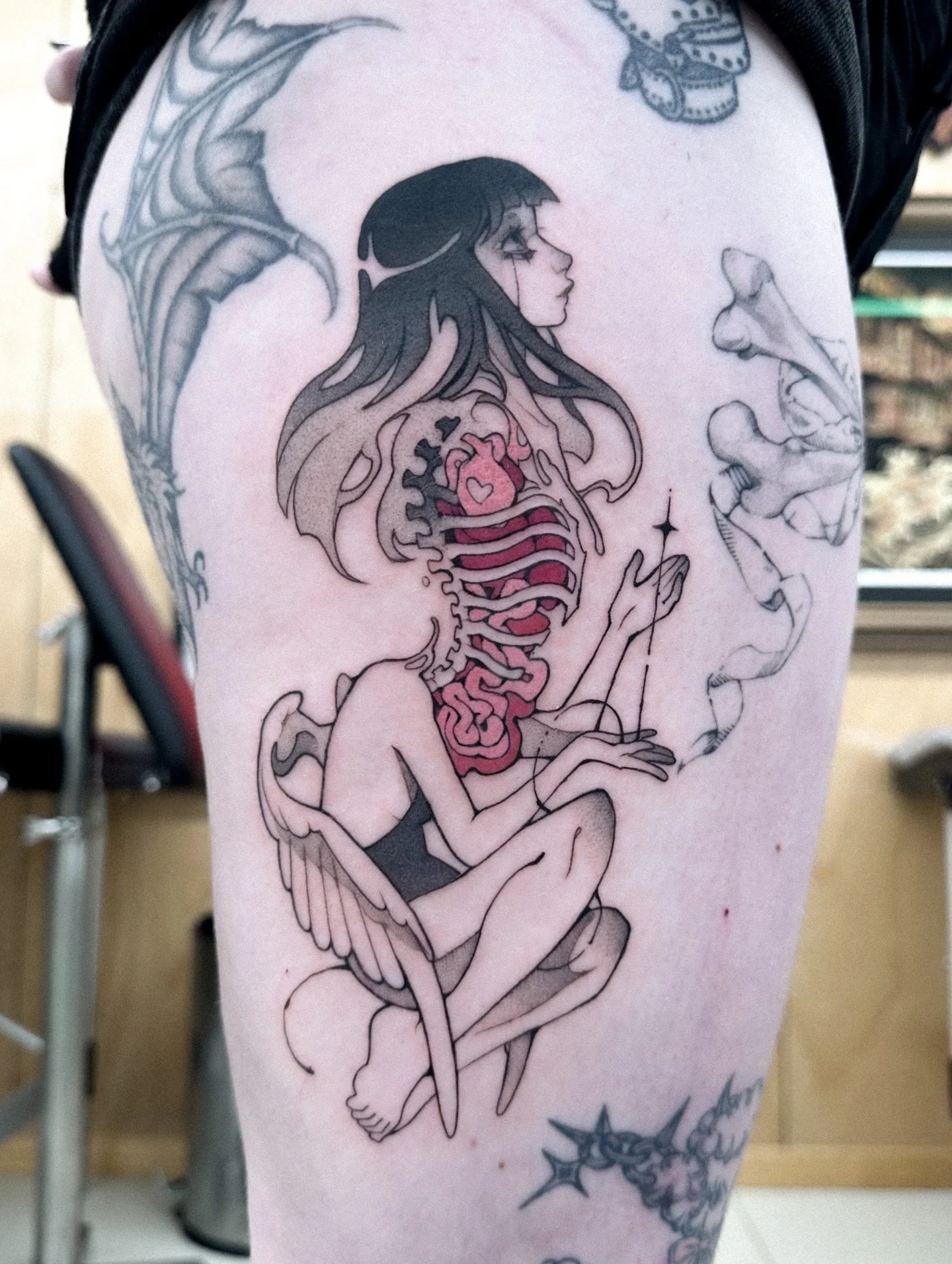 A tattoo of a woman with long hair, sitting cross-legged, holding a needle and thread, with a human heart inside her ribcage, surrounded by decorative elements like stars and flowers.