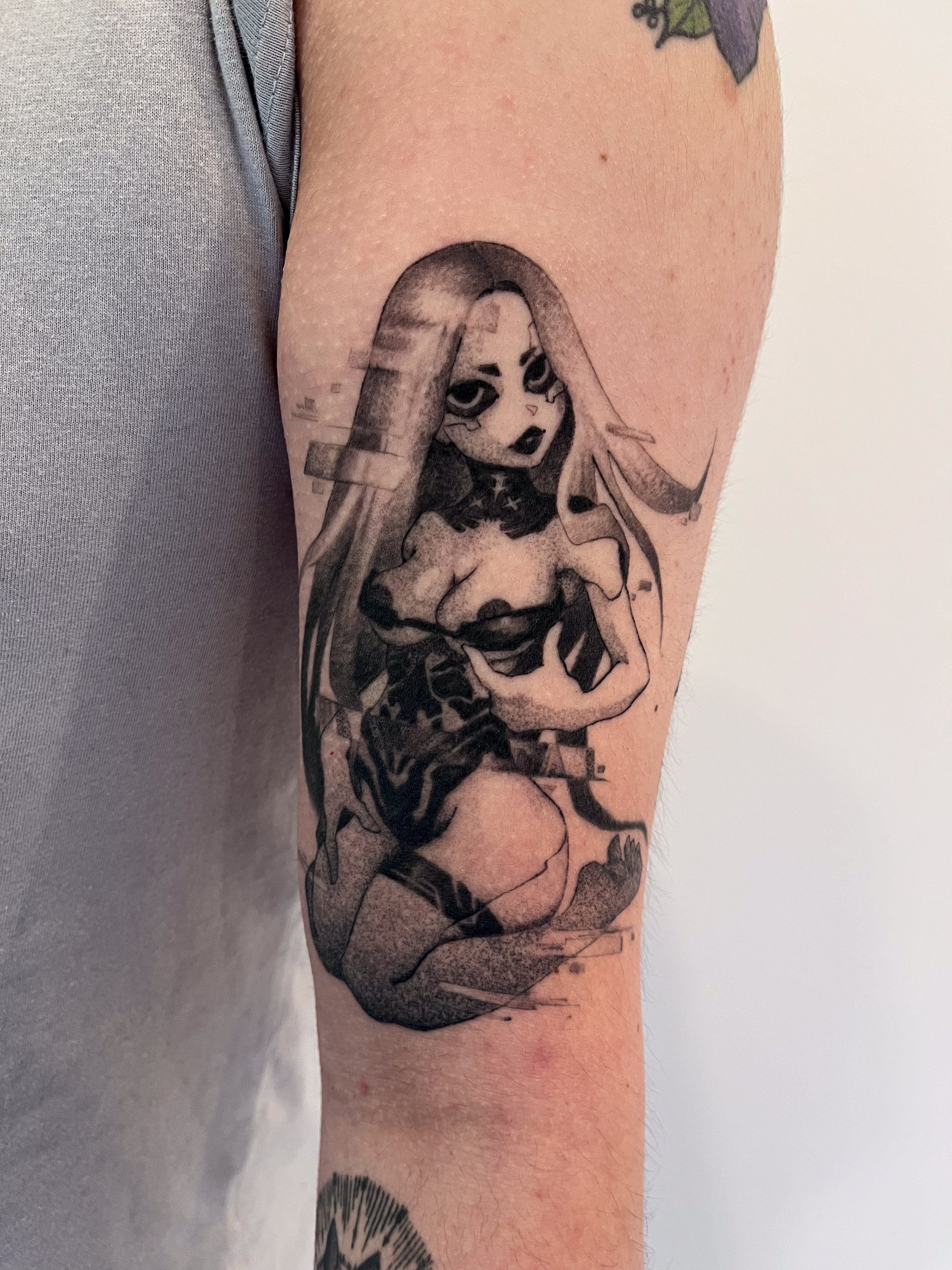 Tattoo of a stylized female character with long hair, dark makeup, and a serious expression, holding a weapon, on a person's upper arm.