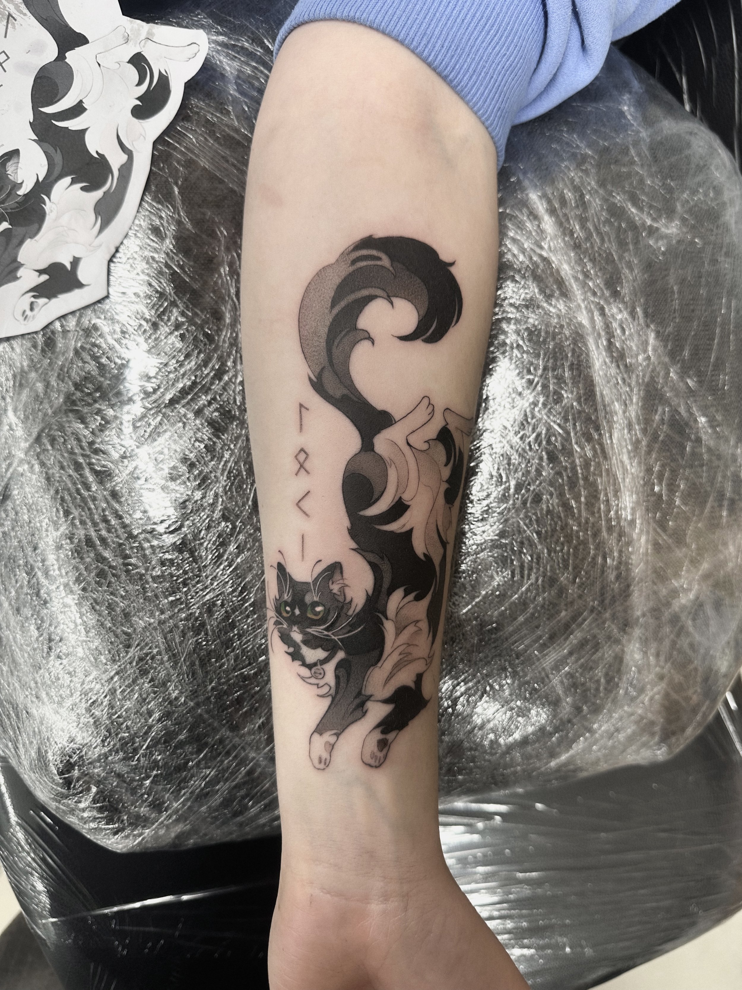 Newly tattooed black and gray cat with piercing green eyes on a person's forearm, with decorative waves and abstract elements surrounding the cat.
