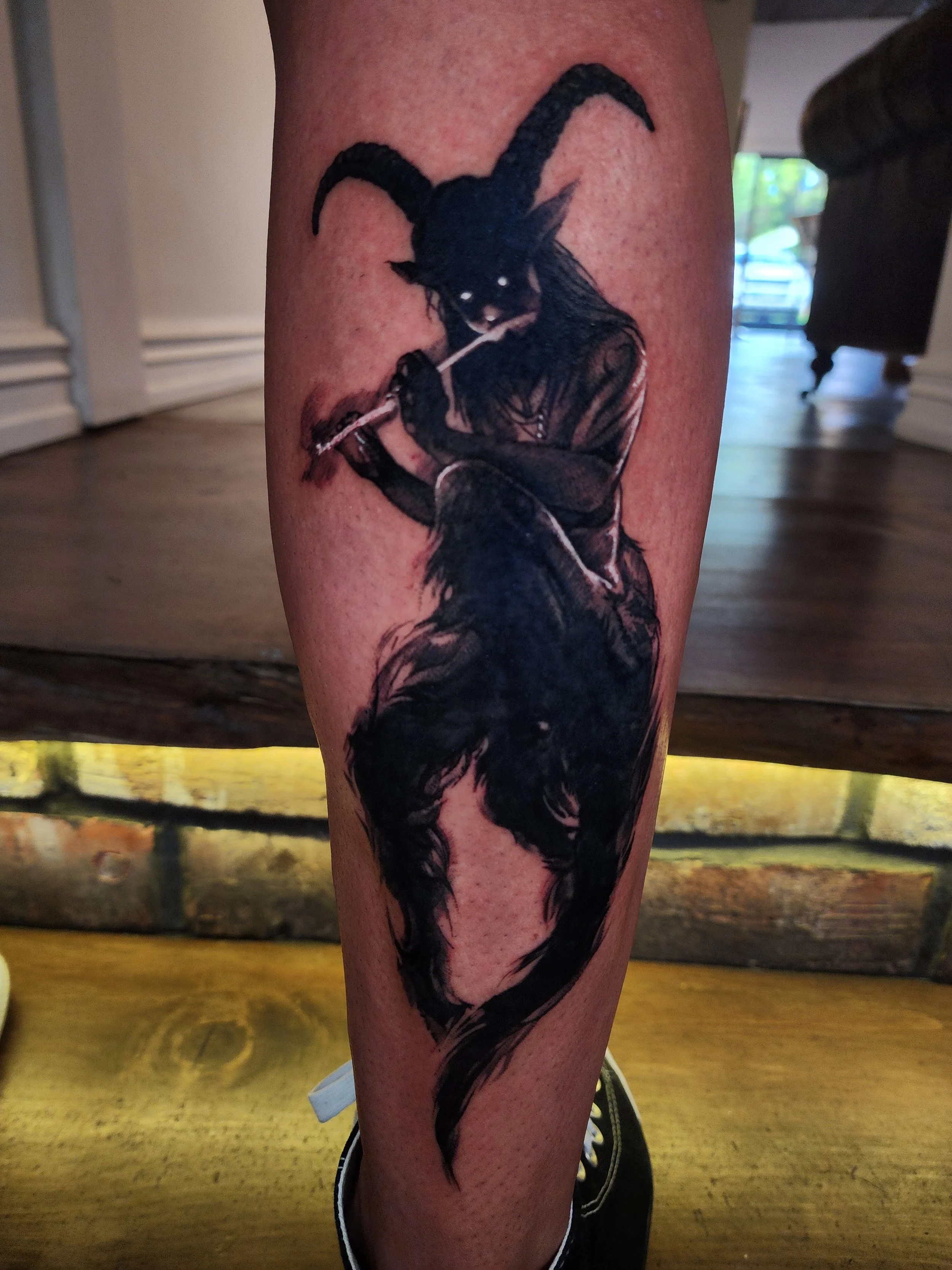 Tattoo of a mythical creature with the body of a dog, the head of a goat, large curved horns, and playing a flute, on a person's calf.