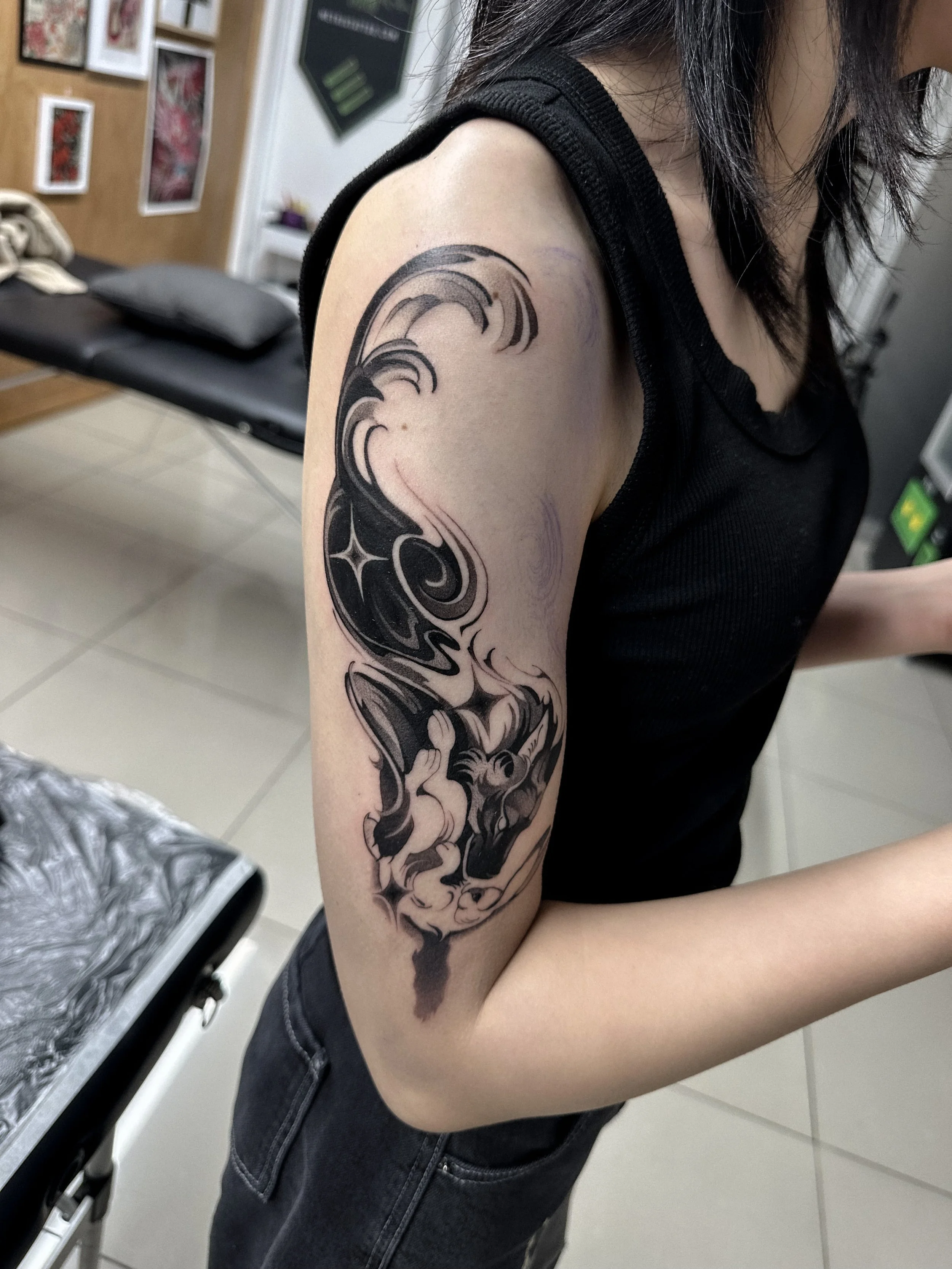 A person with black hair wearing a black sleeveless top has a large black and gray tattoo of a wave on their upper arm.